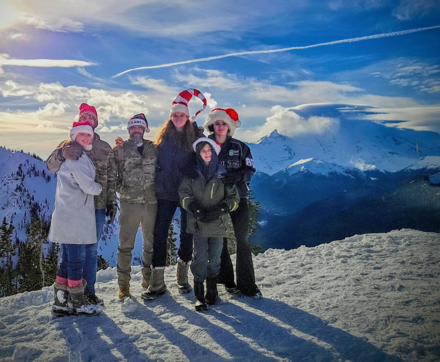 A group wearing winter gear and Santa hats poses together on a snowy mountain peak with scenic views in the background.