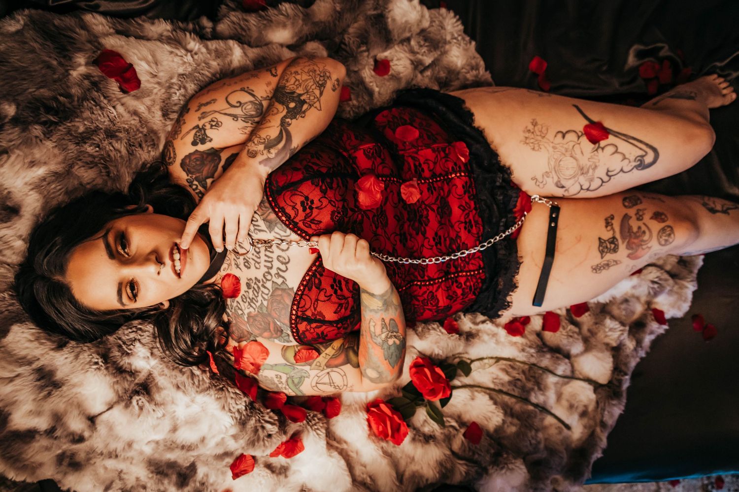 Tattooed woman in red lace lingerie lying on fur with rose petals, holding a decorative pillow.