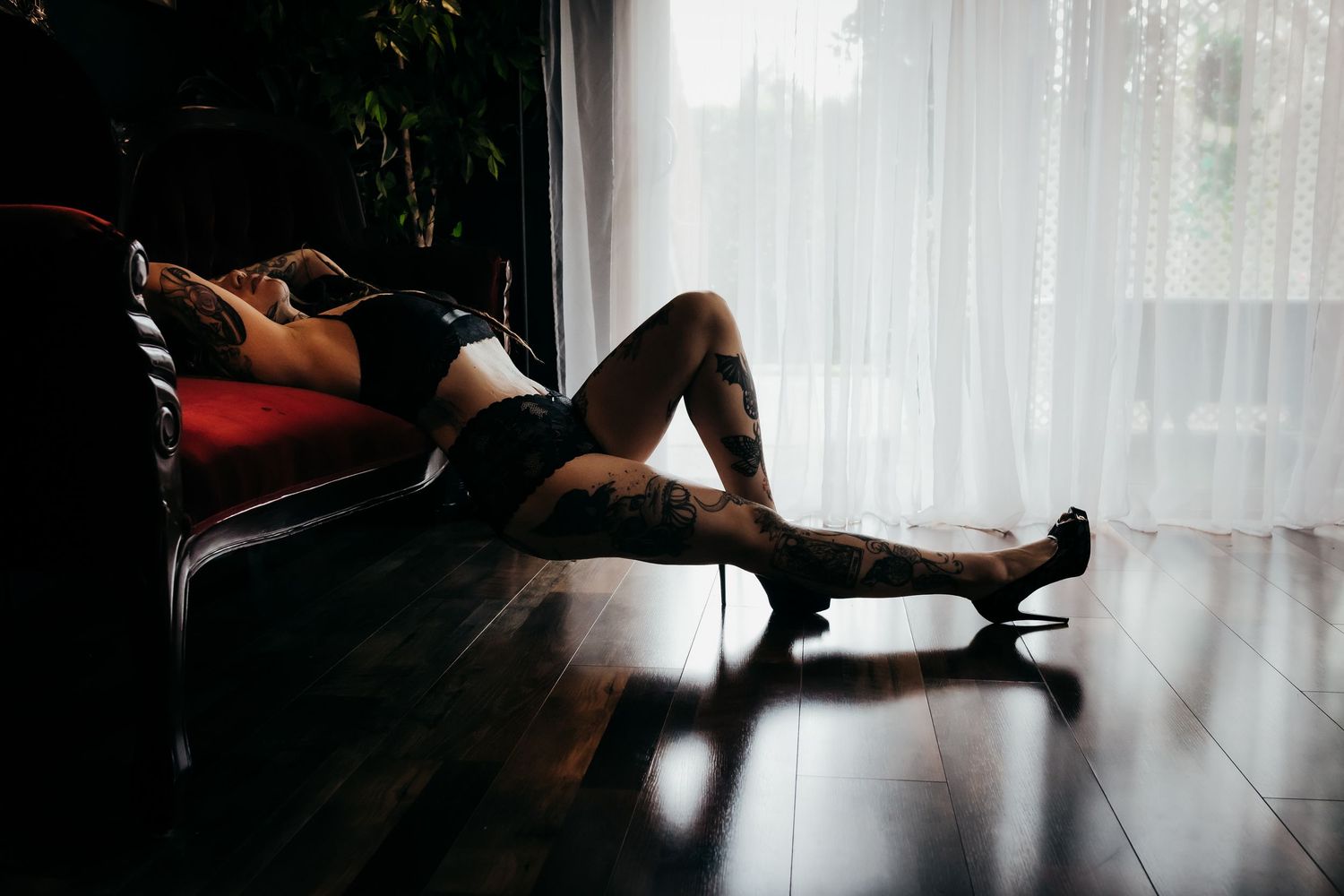 A tattooed woman in lingerie reclines on a vintage sofa near sheer curtains in moody boudoir photography.