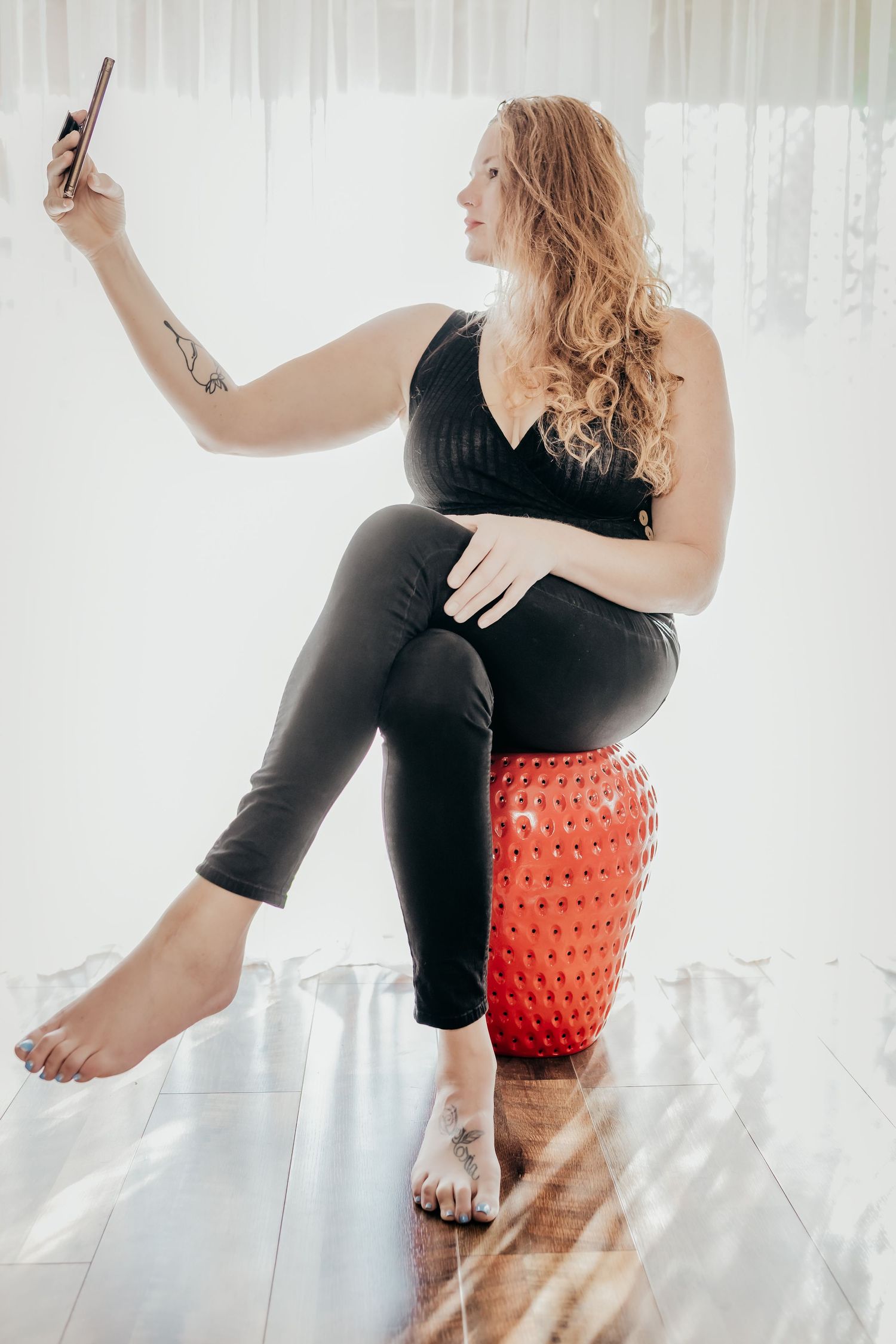 Woman with curly red hair sits on red stool taking selfie, wearing black outfit, showing tattoos, barefoot indoors.
