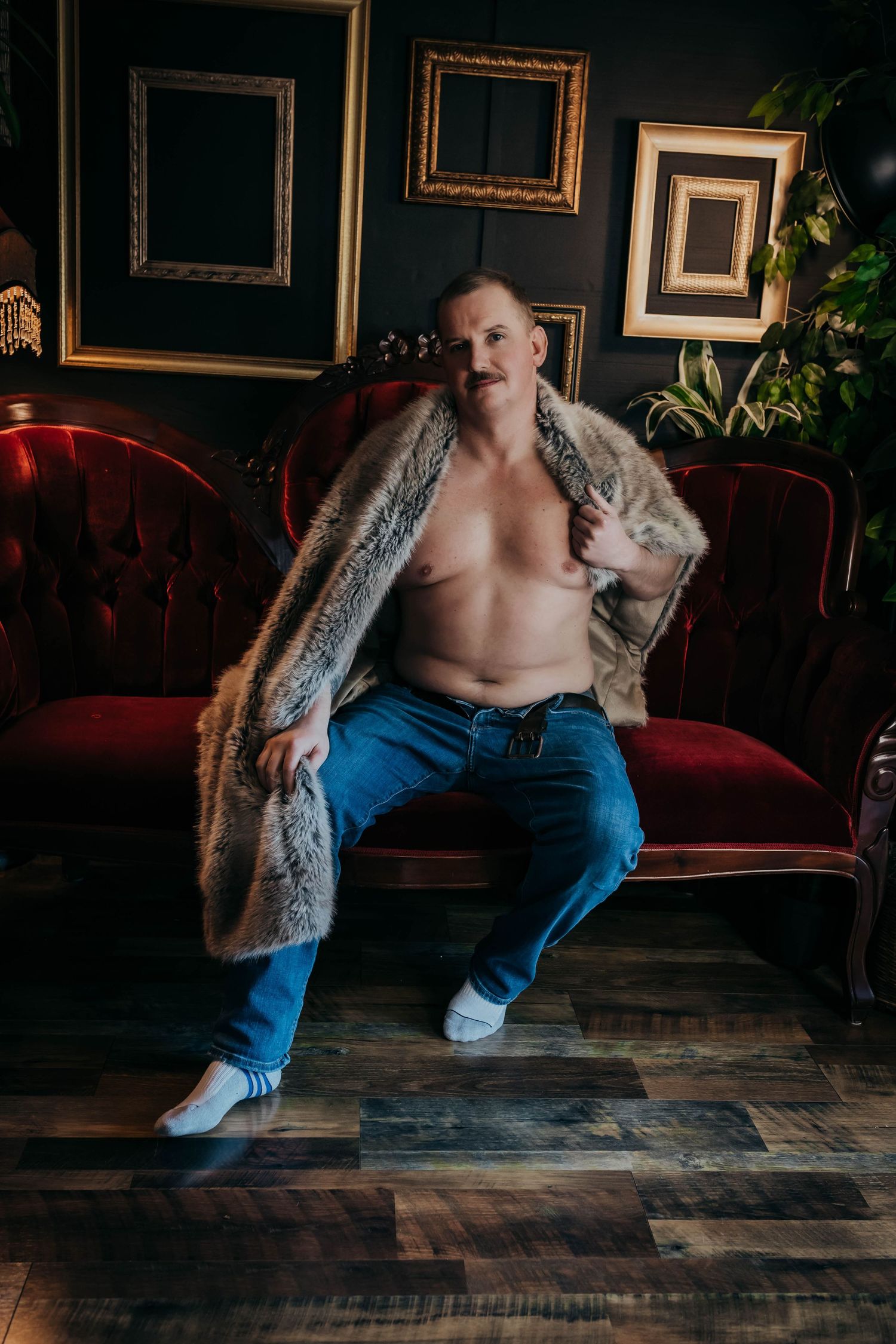 Shirtless man in fur coat and jeans sits on red velvet sofa with empty gold frames on dark wall.