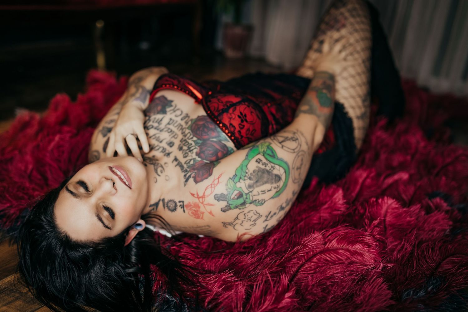 Tattooed woman in red lace lingerie lying on a red fur rug, showcasing extensive body art.