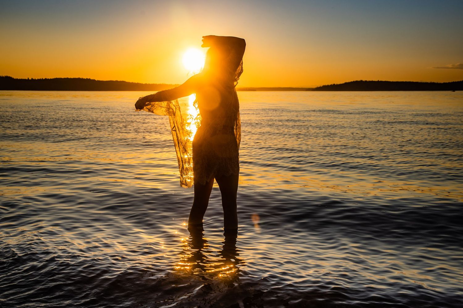 Silhouette of a person standing in shallow water holding a fishing net against a vibrant golden sunset over calm ocean waters.