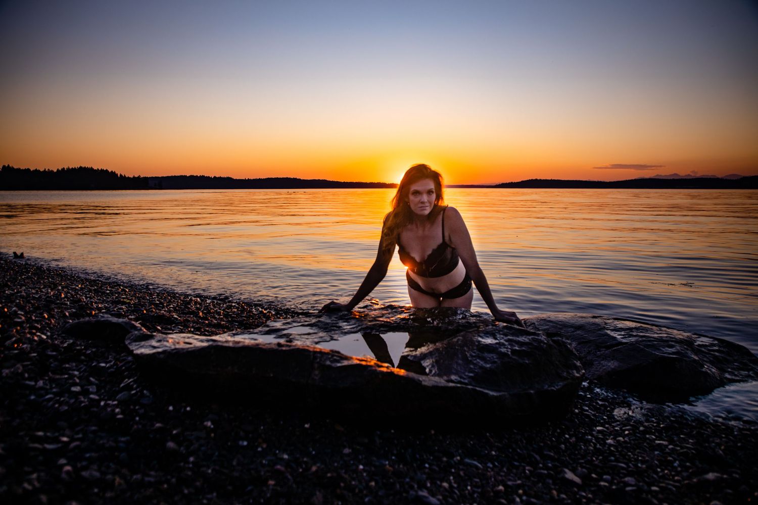 Woman in bikini posing on rocky shore at golden sunset with calm water reflecting warm orange hues.