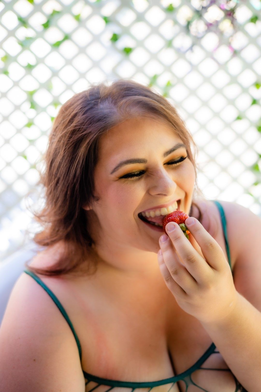 Young woman with bold eye makeup smiling while eating a fresh strawberry outdoors near a lattice fence.