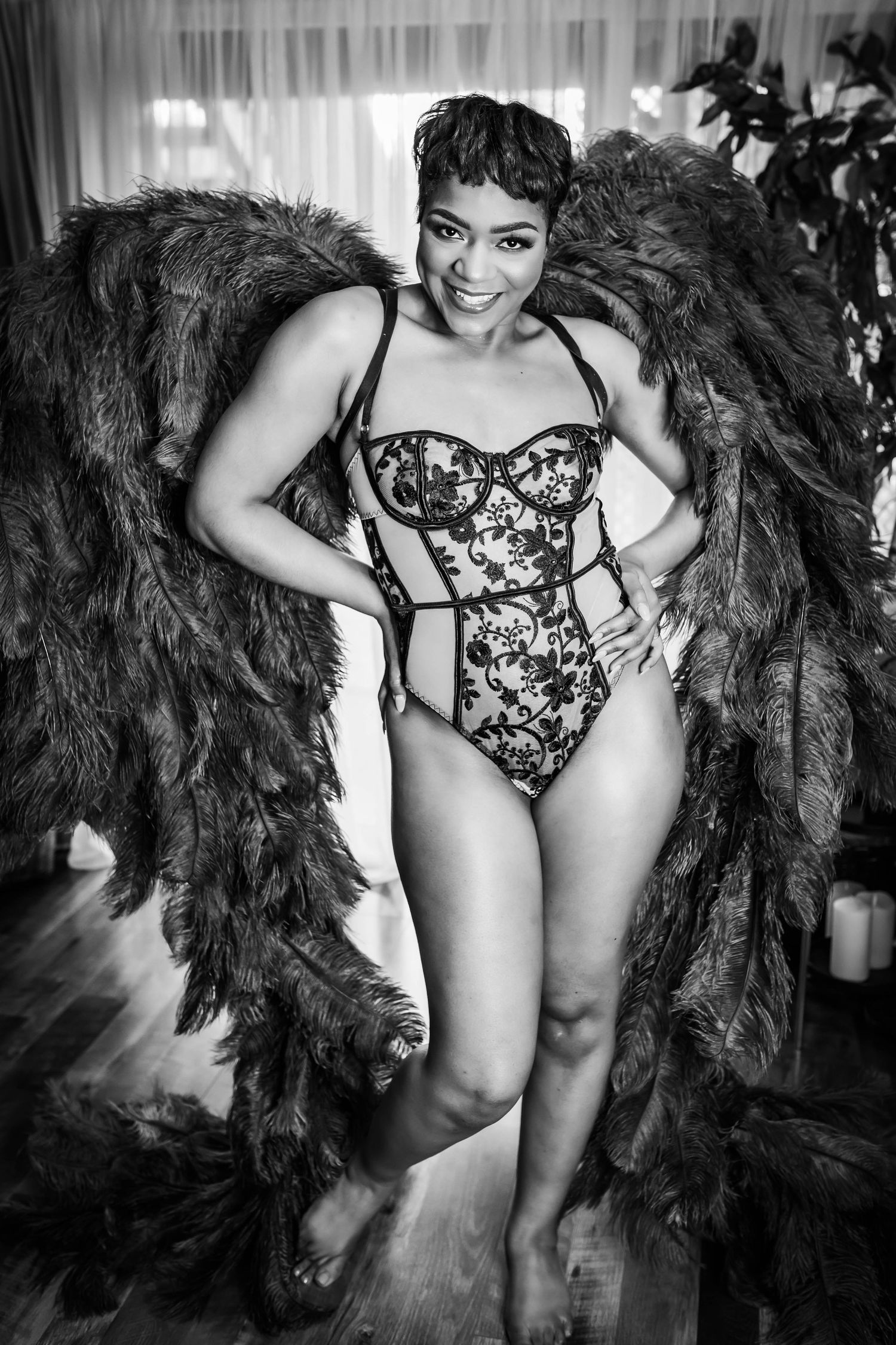 Black and white photo of a smiling woman in lace bodysuit with large feather wings, posing indoors.