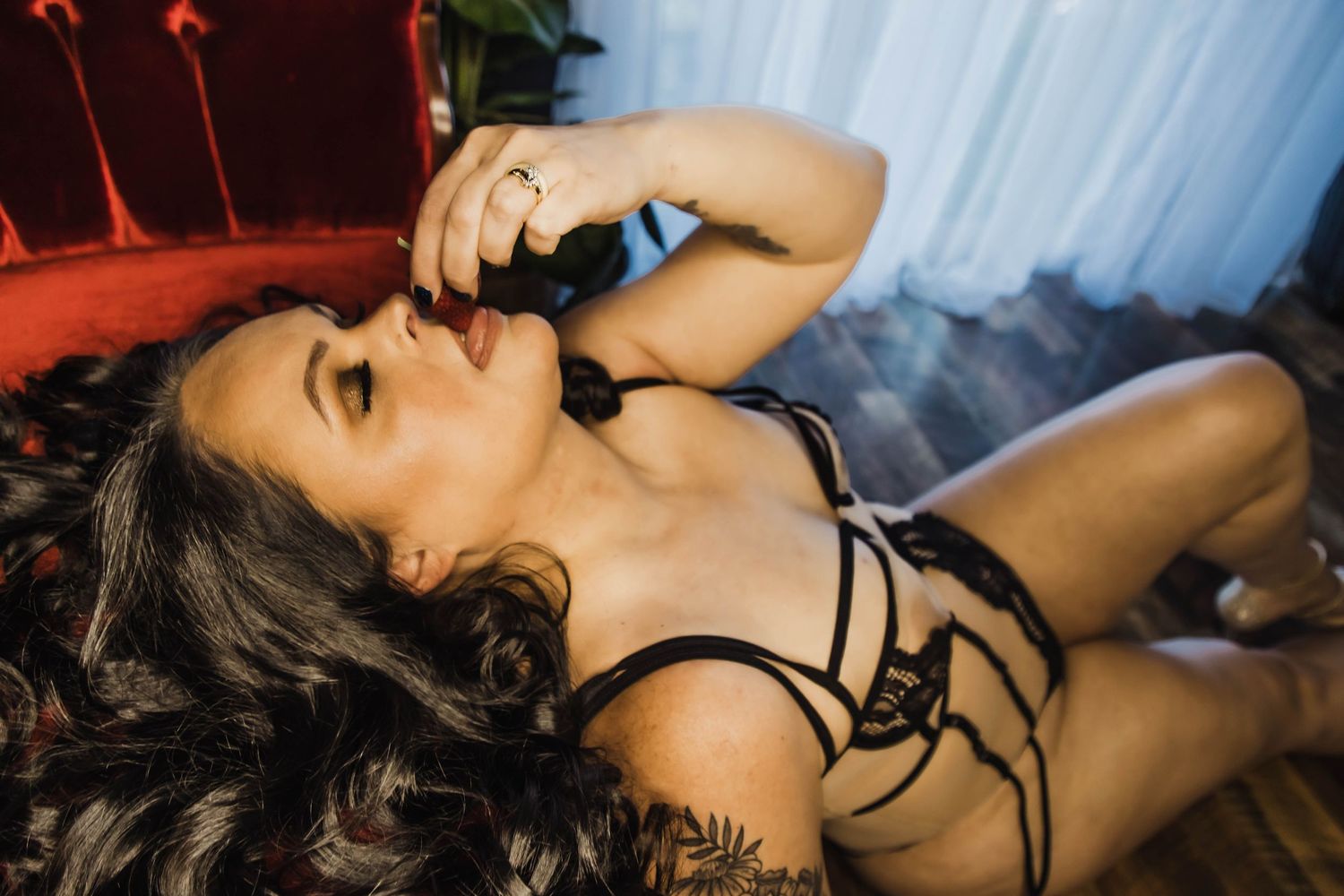 Woman in black lingerie lying on red velvet surface, holding a bottle near her lips in a moody boudoir setting.