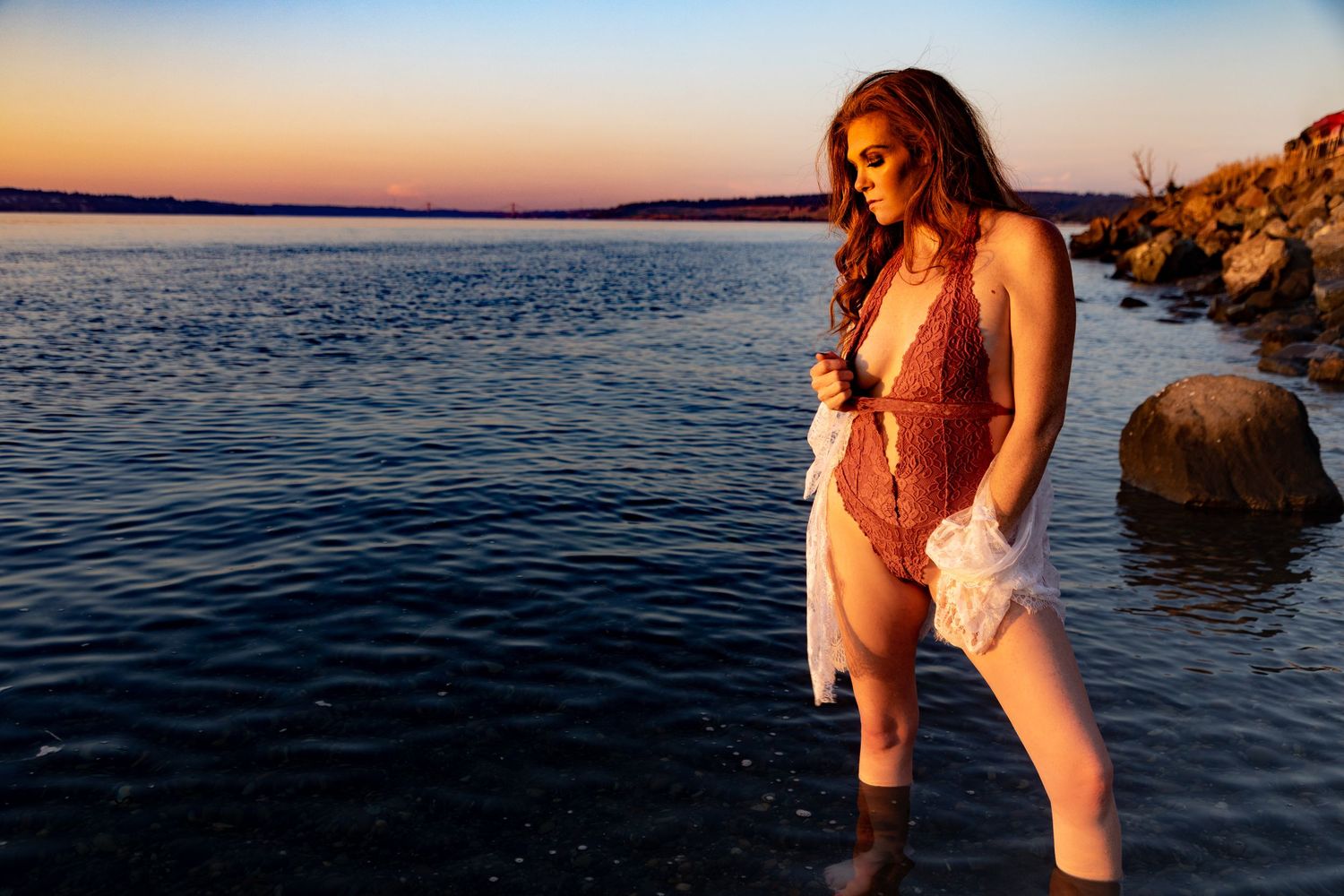 Woman in red lace swimsuit standing in shallow water at sunset with rocky shoreline and colorful sky.