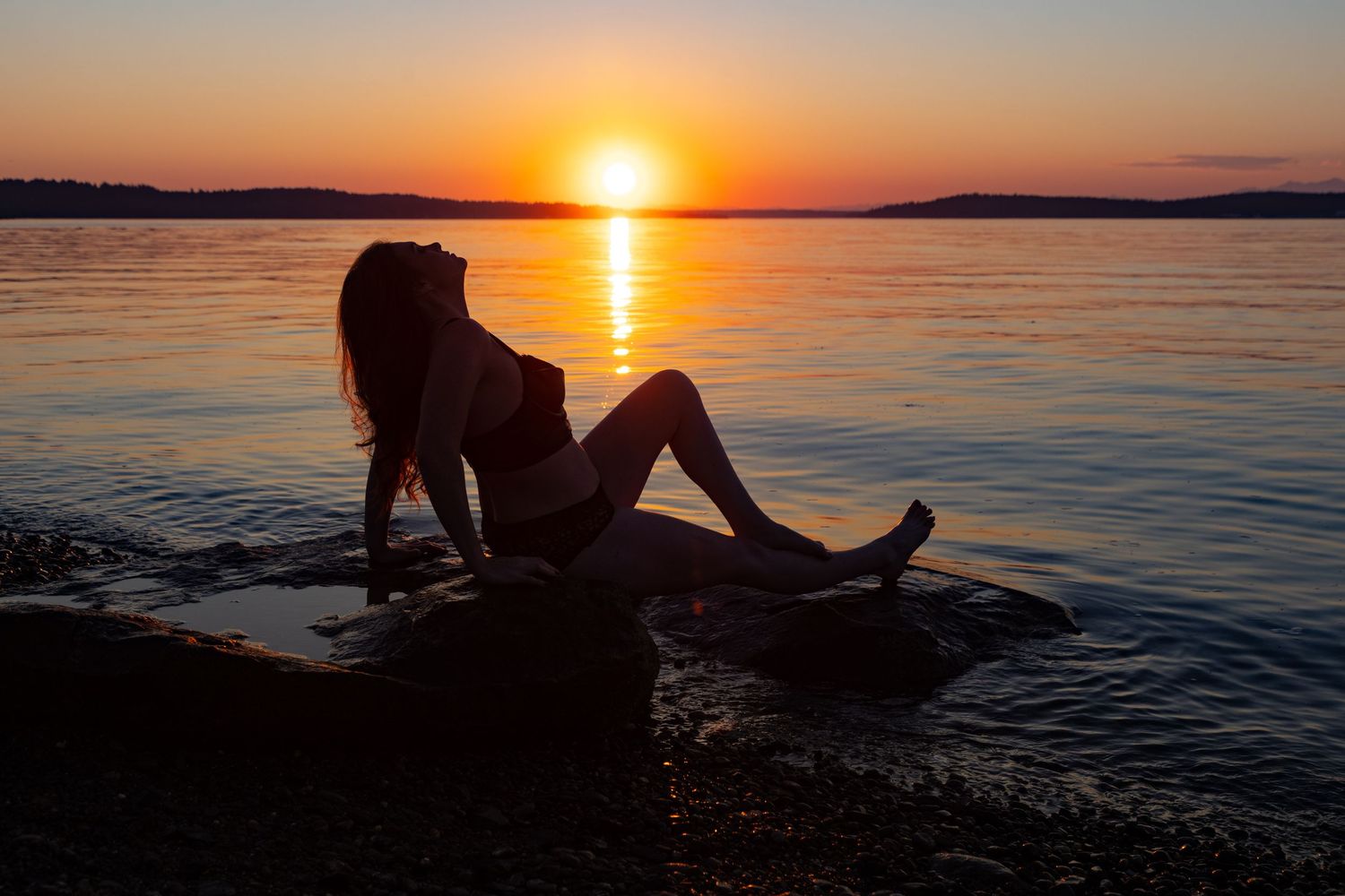 Silhouette of a woman relaxing on rocks by the water during a stunning golden sunset over a calm lake.