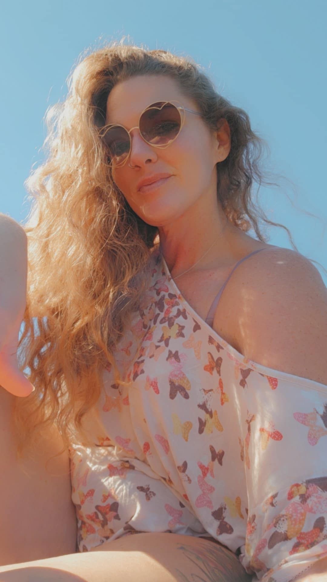 A person with long curly blonde hair wearing round sunglasses and a butterfly-print top poses in warm summer sunlight.