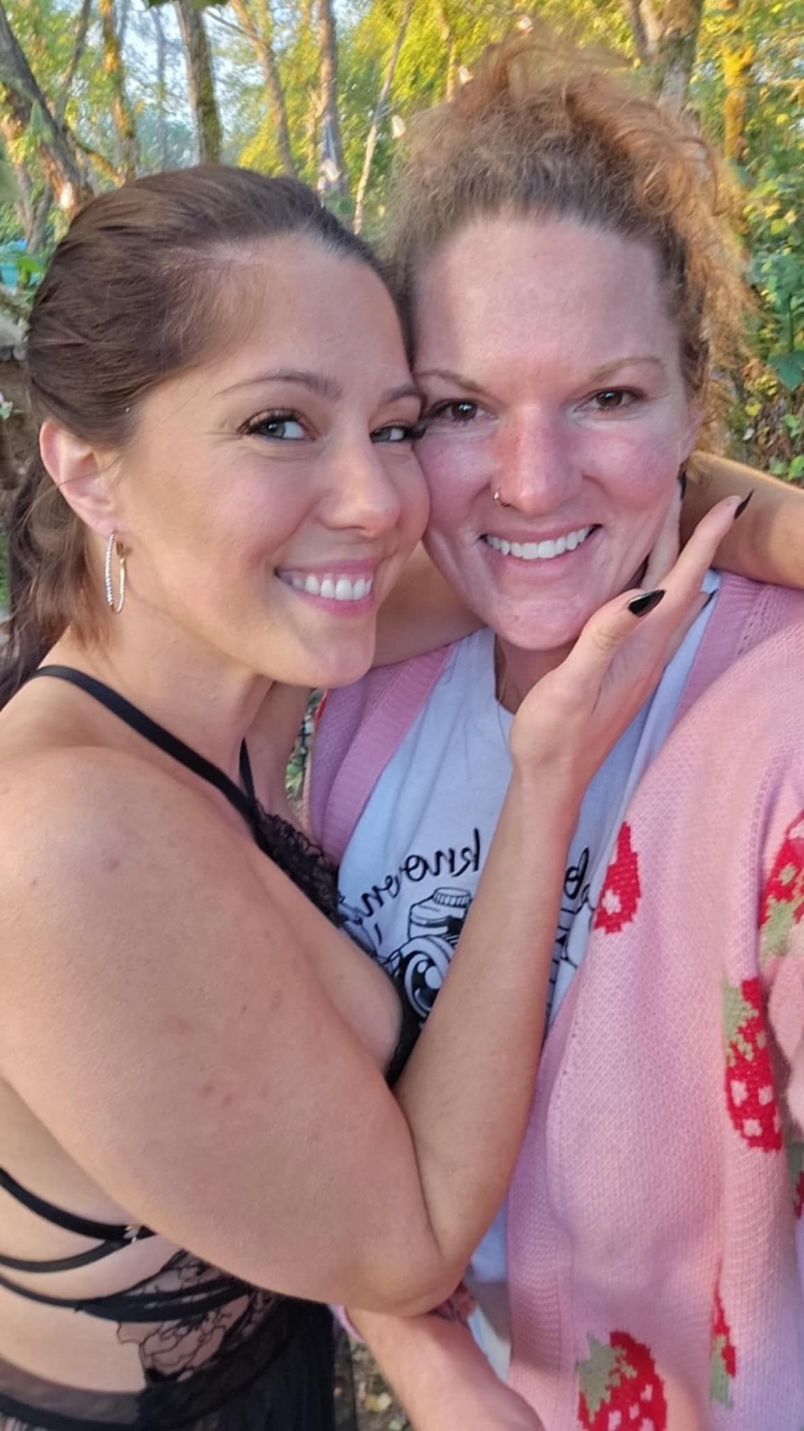Two friends share a close selfie outdoors in casual attire with natural scenery in the background.