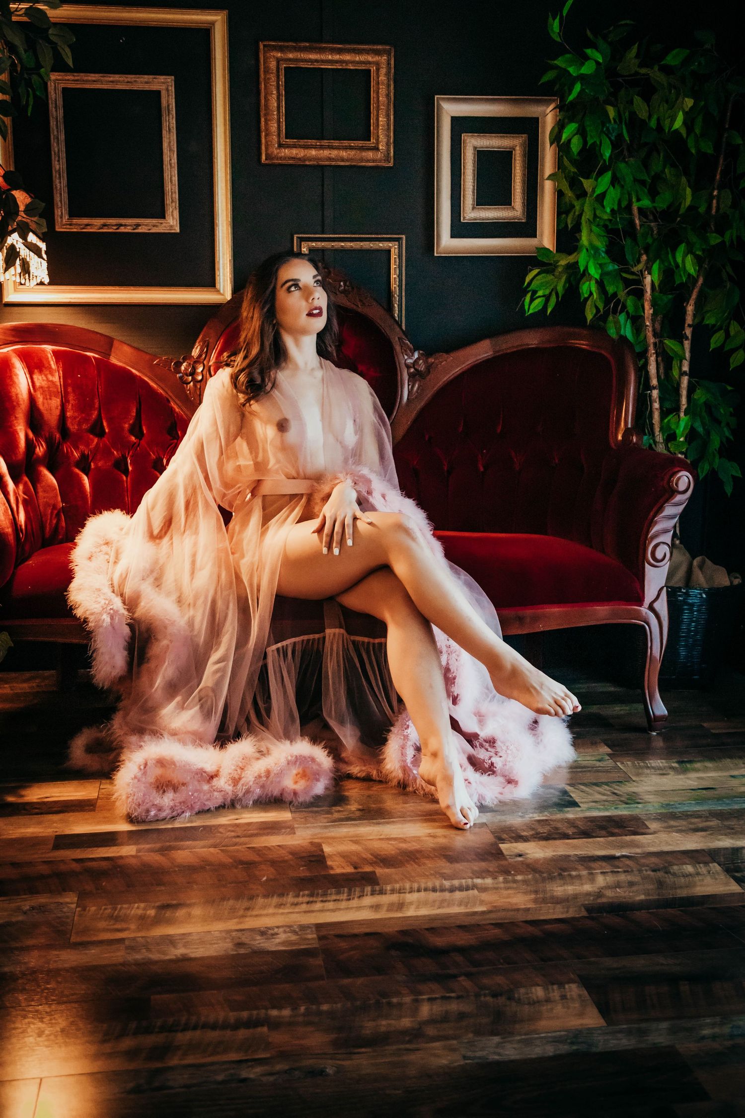 Glamorous boudoir photo featuring peach robe and red velvet couch with empty vintage frames on dark wall.