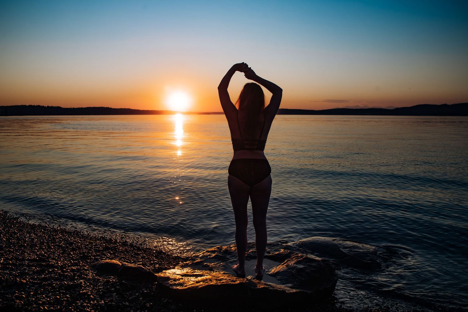 Silhouette of a woman in a bikini posing on rocky beach at golden sunset over calm water.