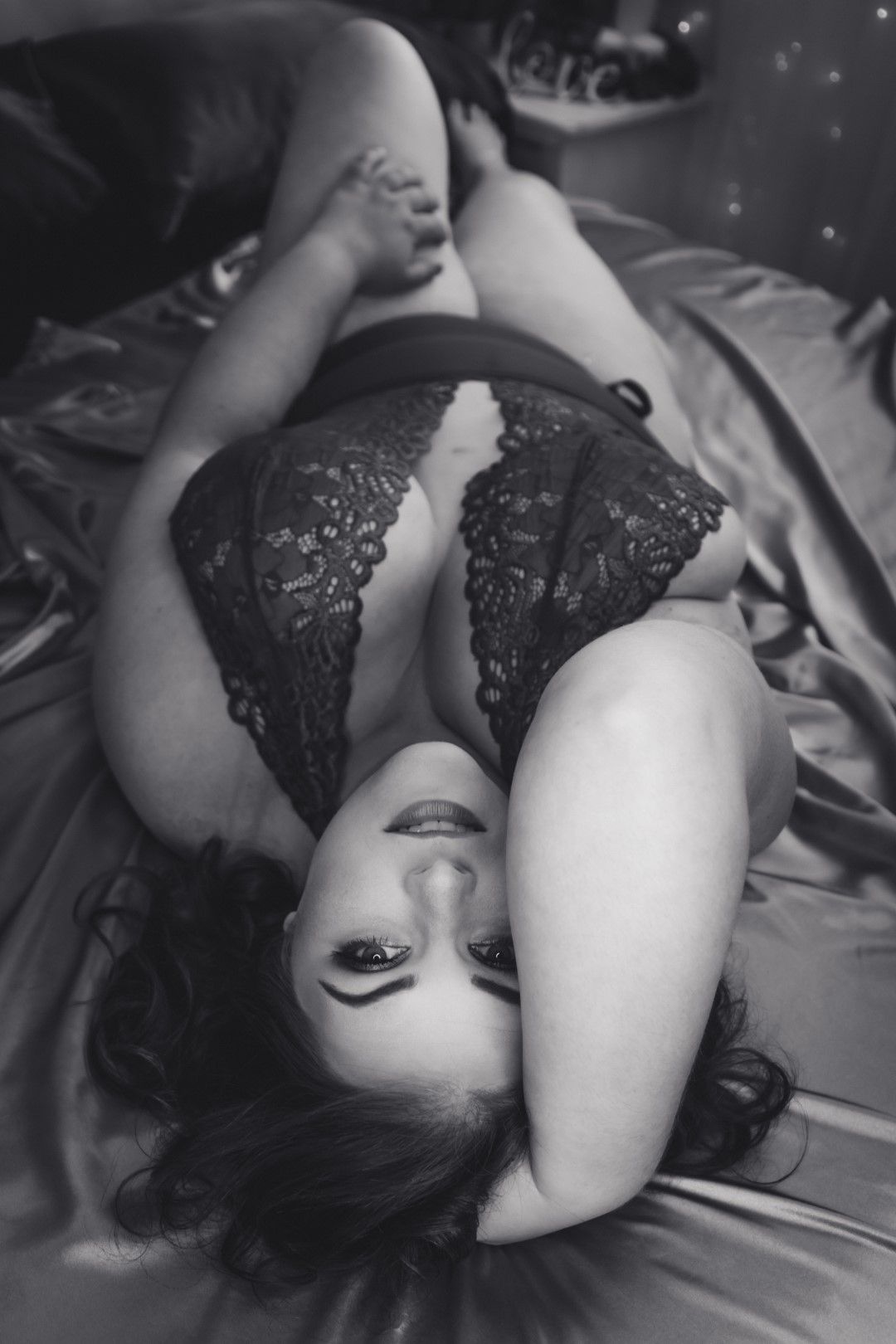 Black and white boudoir photo of a woman in lace lingerie lying on satin sheets.