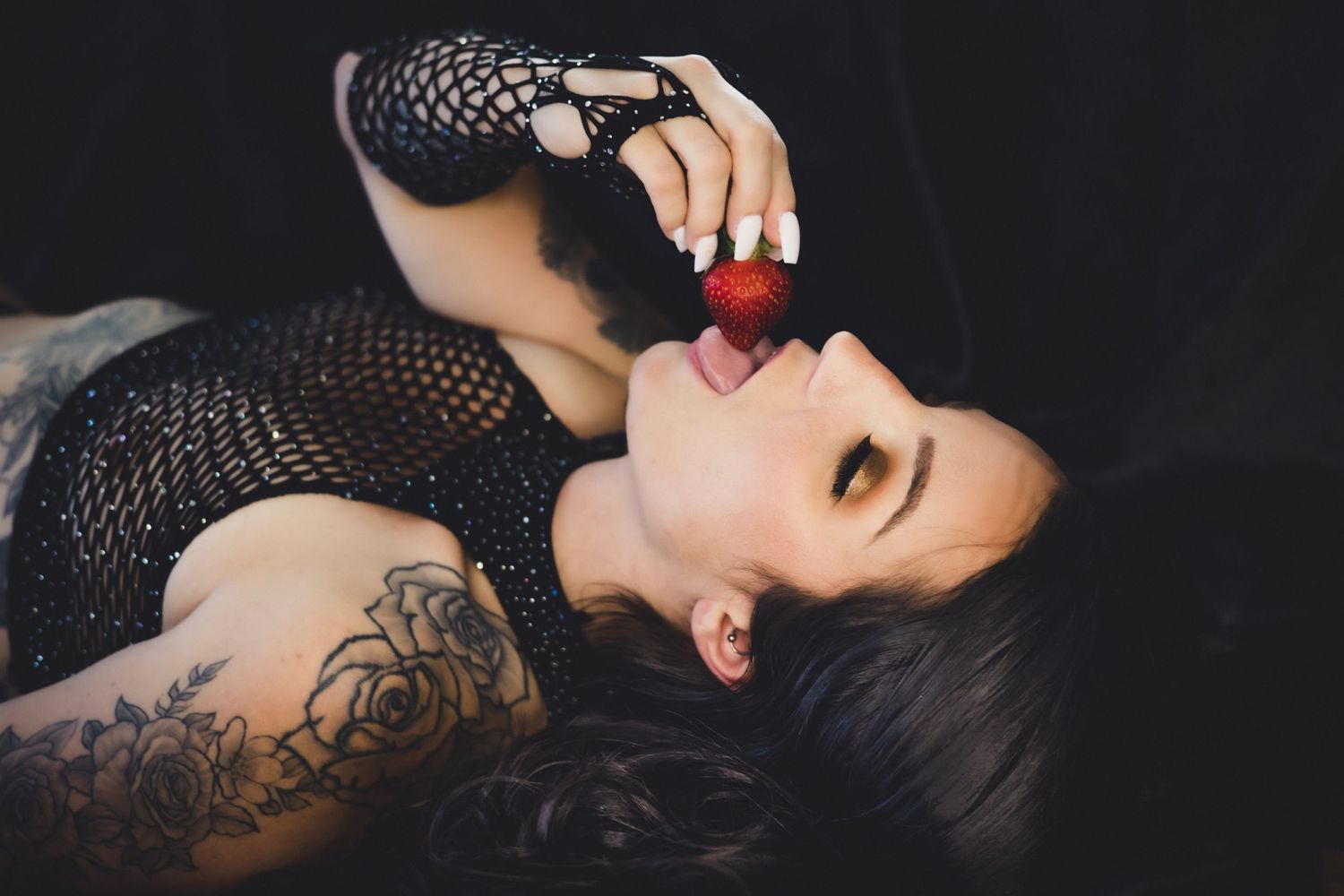 Artistic portrait of a person in black lace with tattoos lying down while holding a red cherry to their lips.