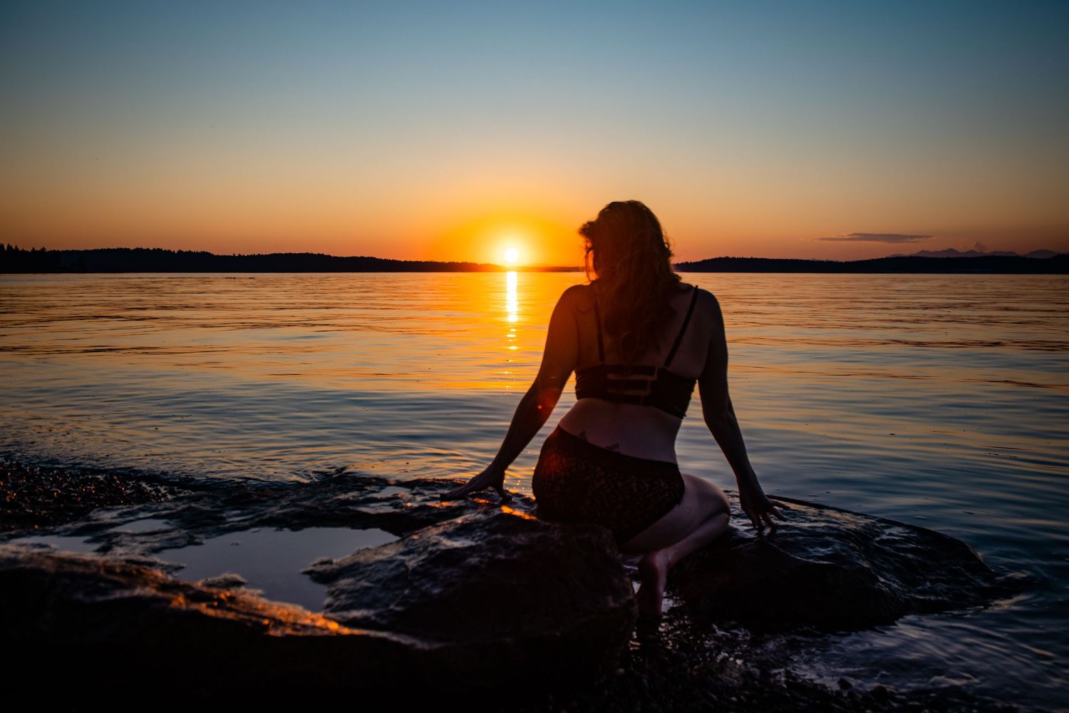Woman sitting on rocks by calm water watching a vibrant golden sunset over the horizon at dusk.