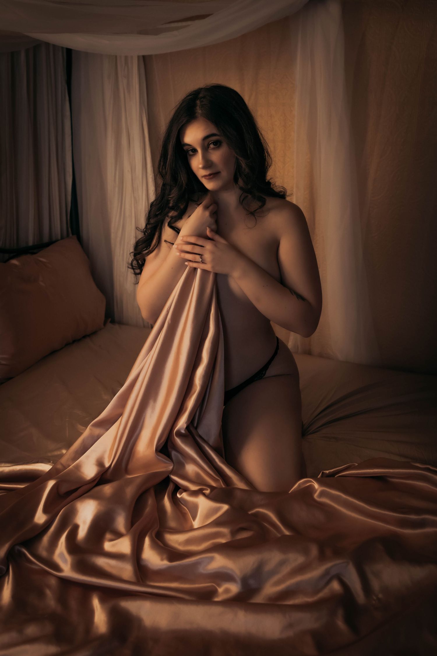 A woman draped in golden satin sheets poses elegantly on a bed in warm, moody boudoir photography lighting.