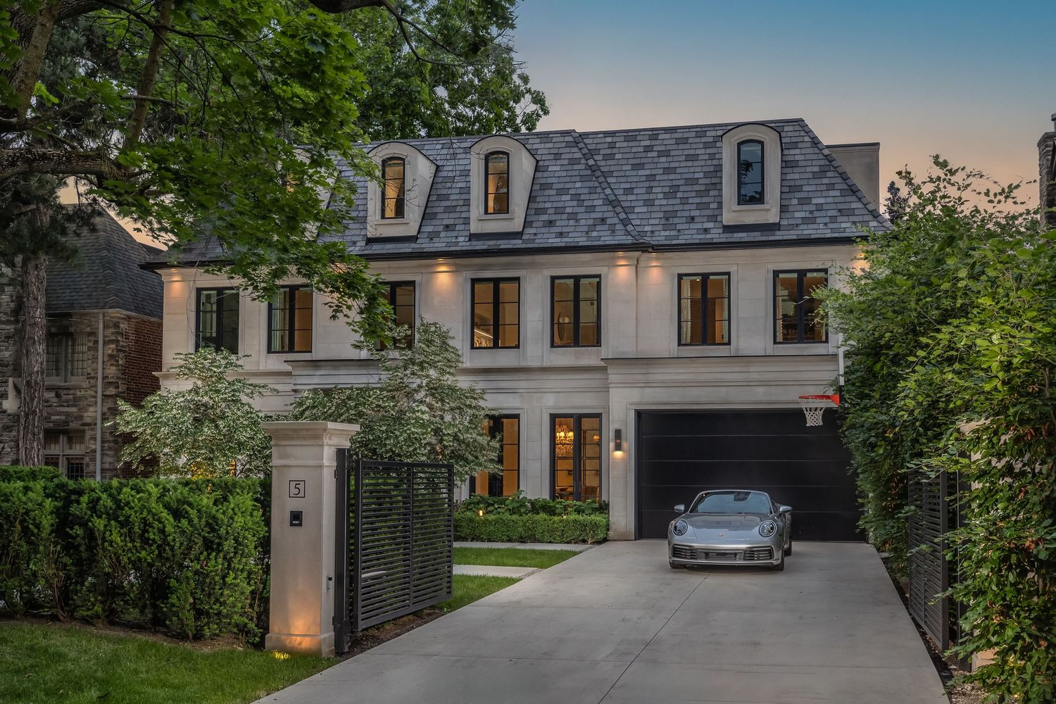 Luxury stone mansion with landscaped driveway and manicured gardens illuminated at dusk against a blue sky.