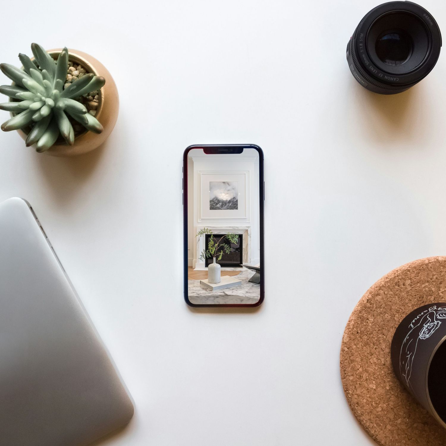 Minimalist desk setup with iPhone, succulent plant, laptop edge, and cork coaster on white surface.