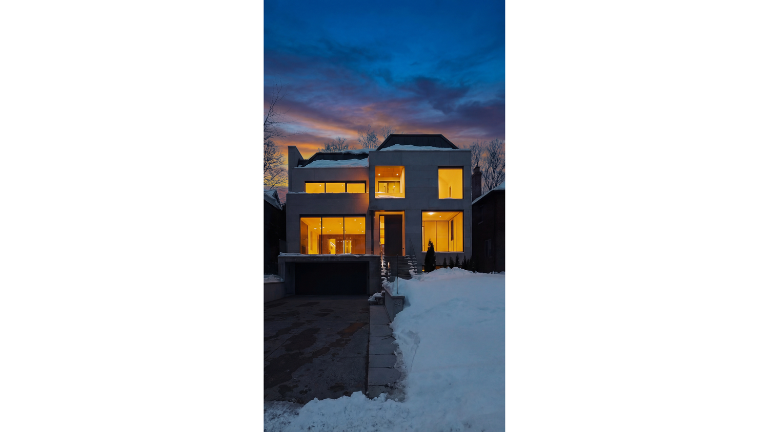 Modern two-story house with warm glowing windows illuminated at dusk against a dramatic blue winter sky and snowy ground.