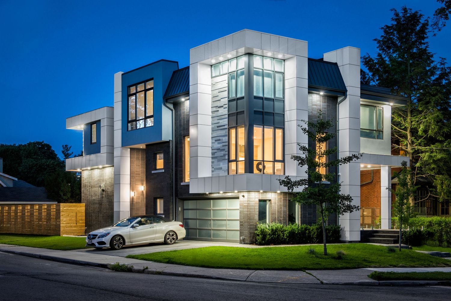 Modern luxury home exterior with white geometric architecture and large windows illuminated at dusk.