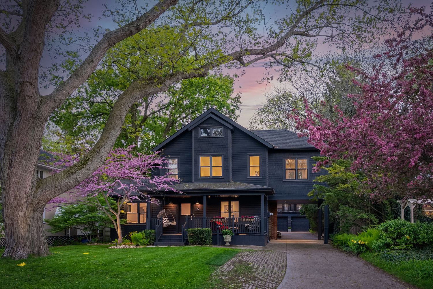 Charming dark blue craftsman house with glowing windows at dusk surrounded by blooming spring trees and a curved driveway.