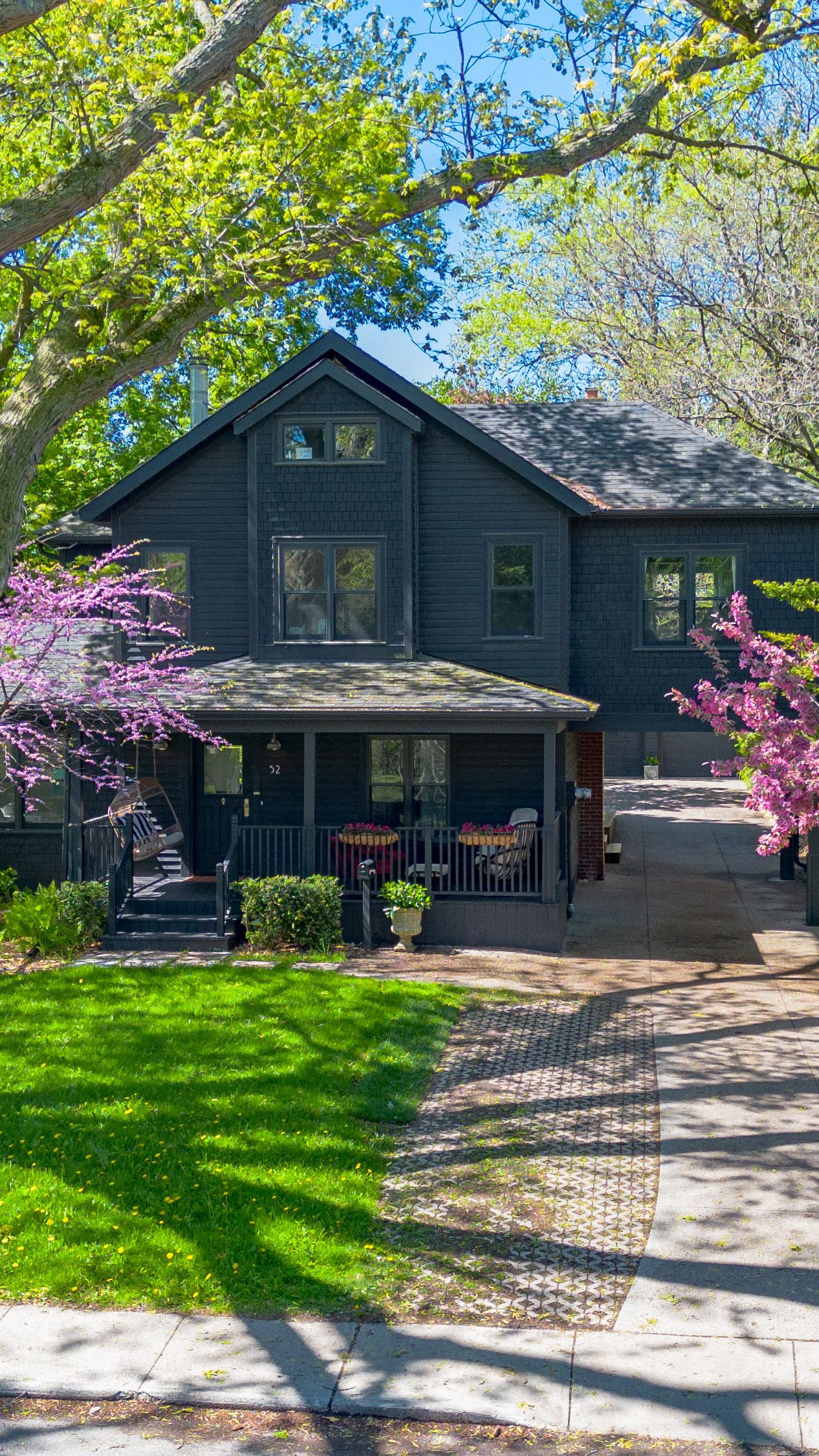 Black wooden ranch house with blooming pink cherry trees and green lawn on a sunny spring day.