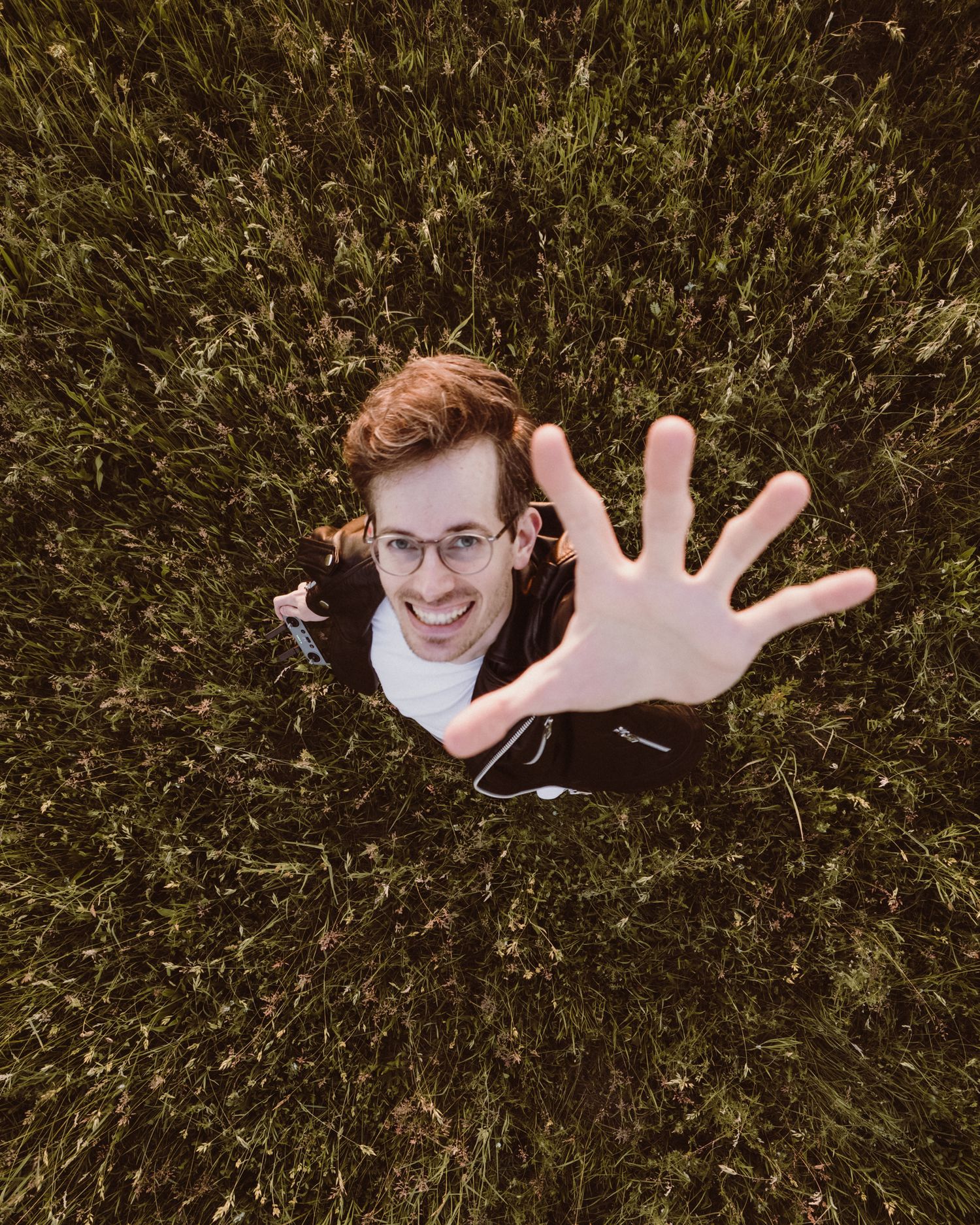 Aerial view looking down at someone laying in grass wearing glasses, reaching upward with one hand toward the camera.