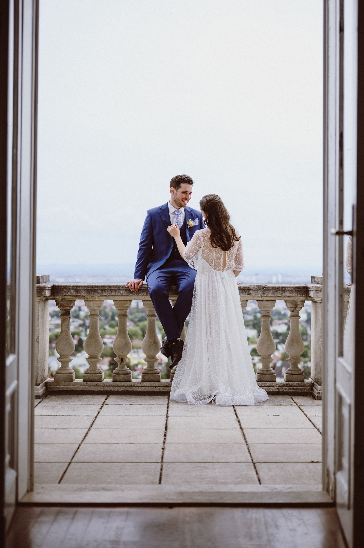 A bride and groom share an intimate moment on a stone balcony overlooking a scenic landscape.
