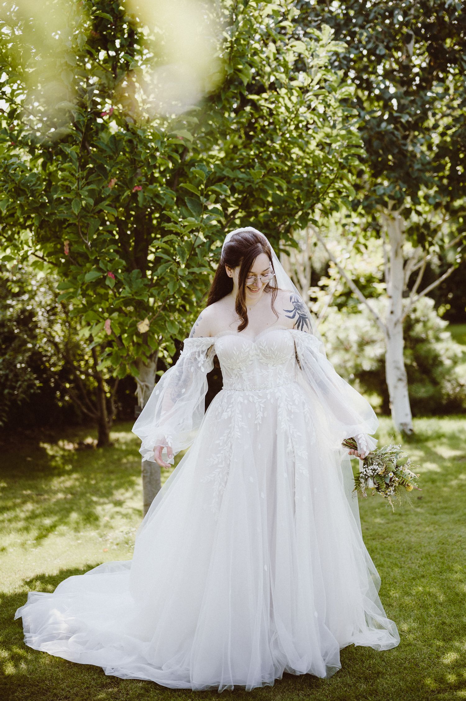 A bride in a flowing white wedding gown twirls gracefully in a sunlit garden setting with trees in the background.
