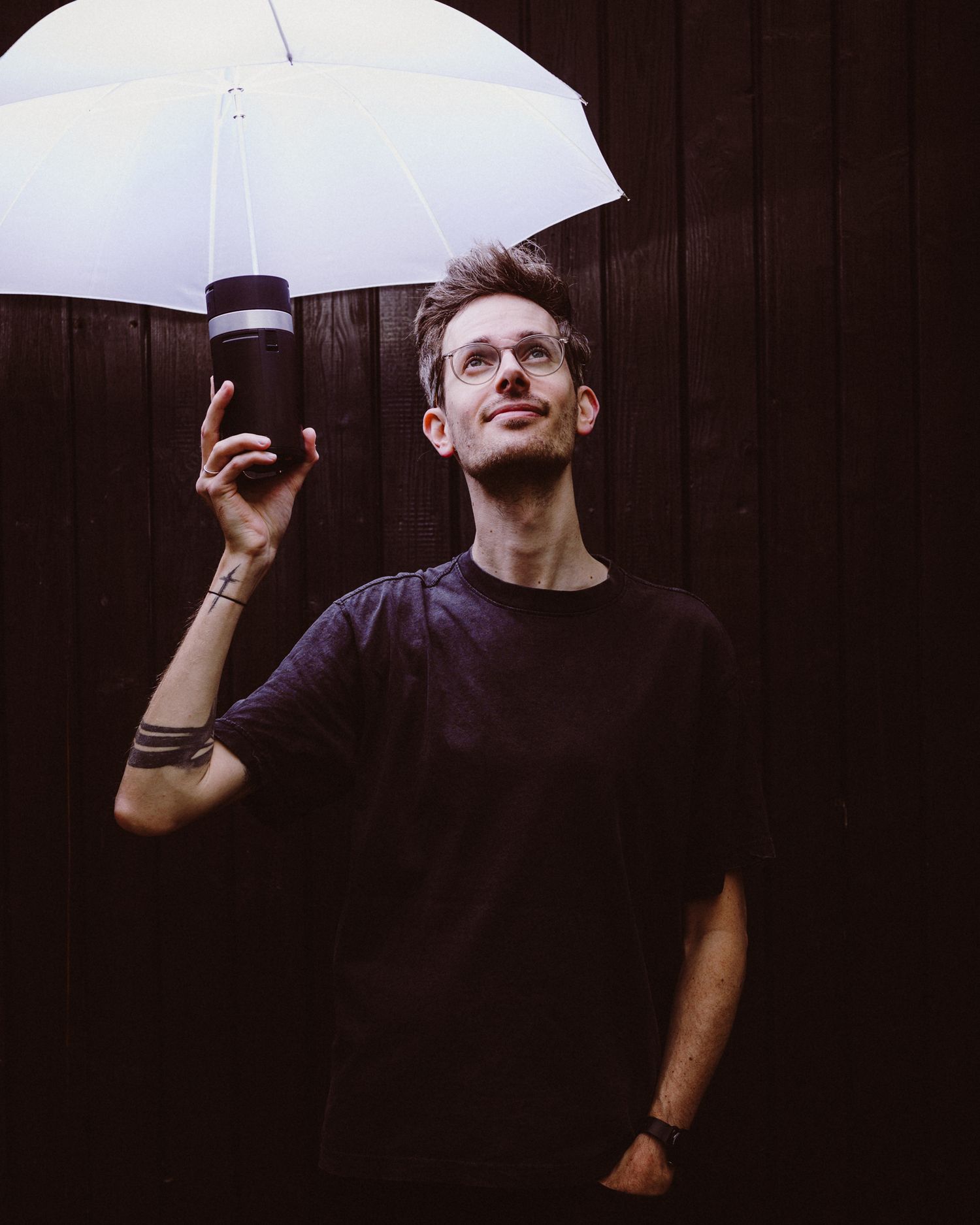Figure holding a coffee cup under a white umbrella against a dark brown wall backdrop in moody lighting.