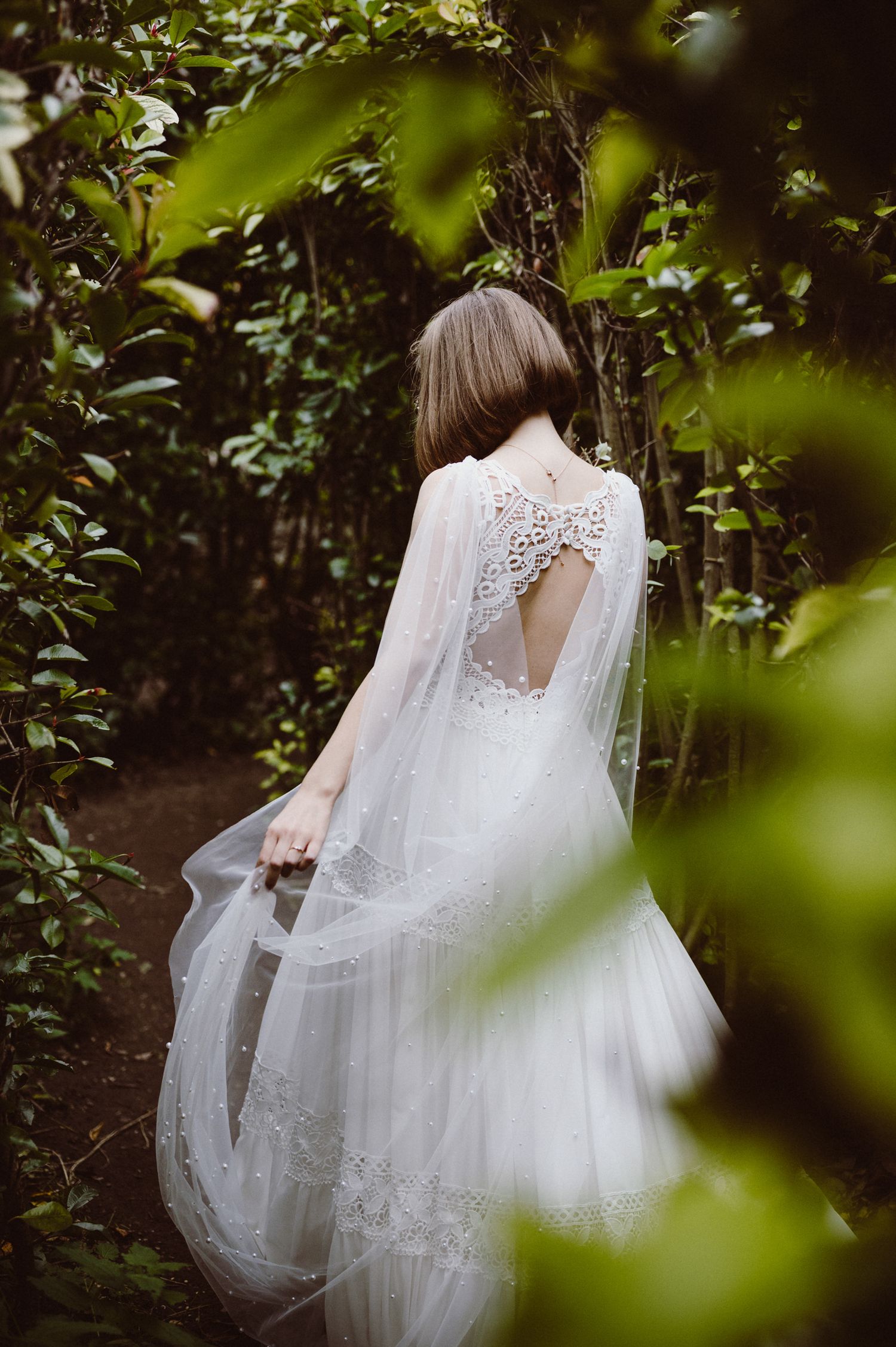 A flowing white wedding dress with delicate lace details and a long veil captured through green foliage in a garden setting.