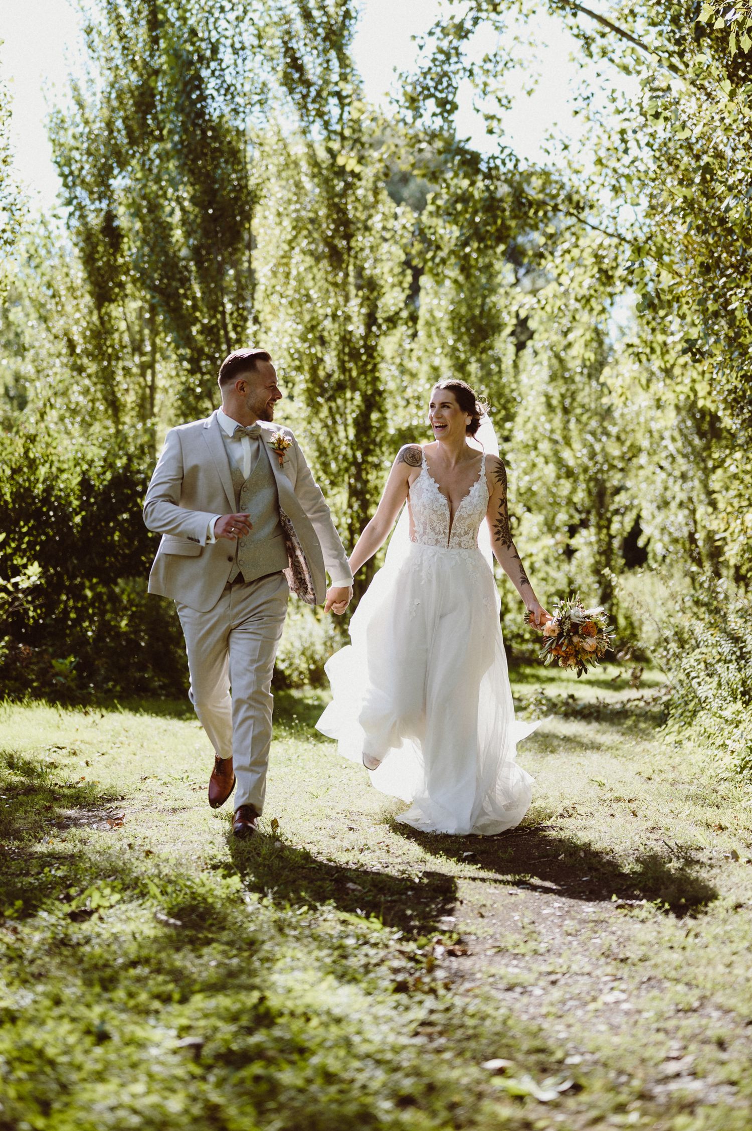 A couple in wedding attire playfully runs and dances together through a sunlit orchard on their special day.