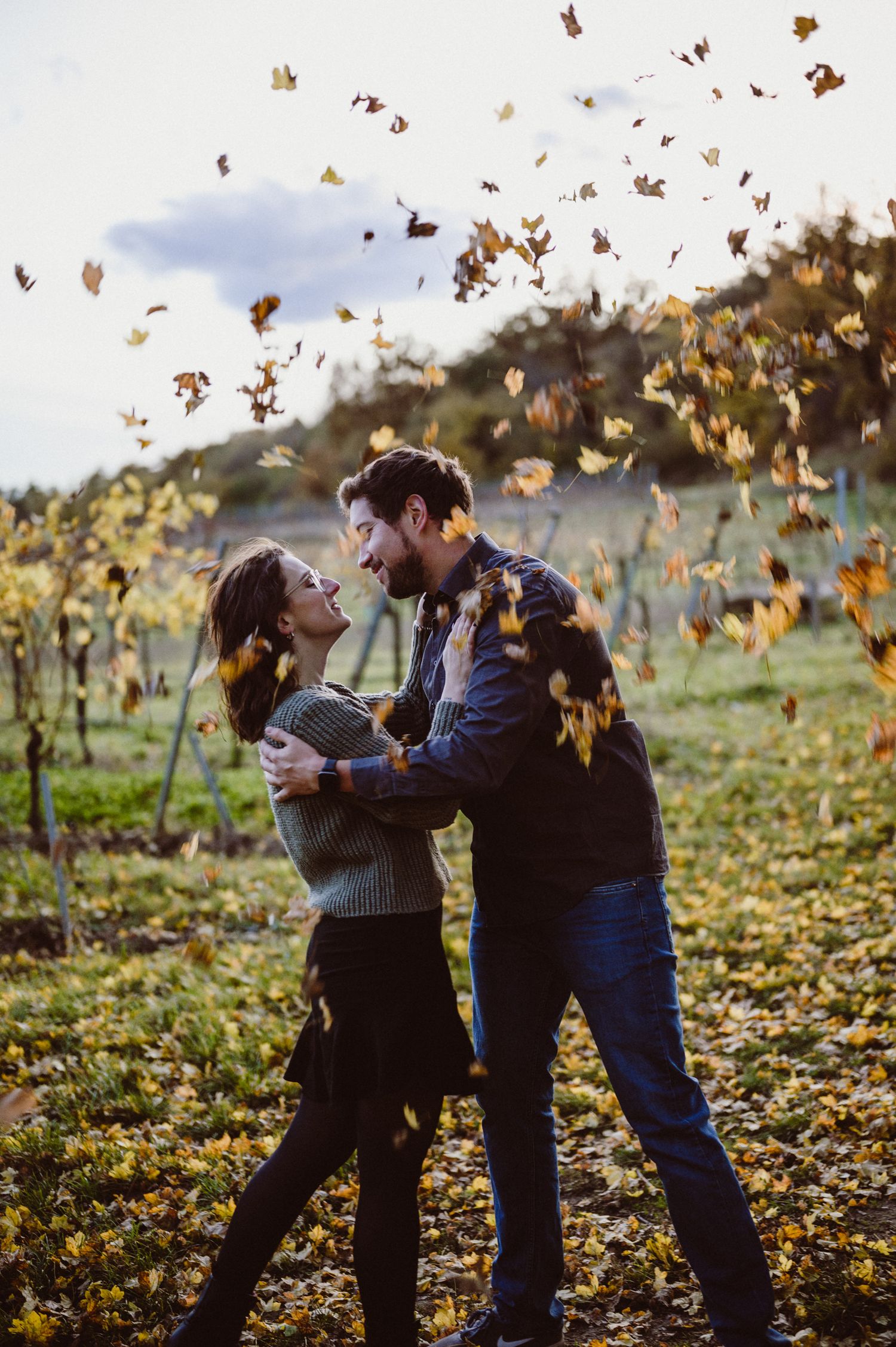 A couple plays together in a field of autumn leaves as golden foliage falls around them in warm sunlight.