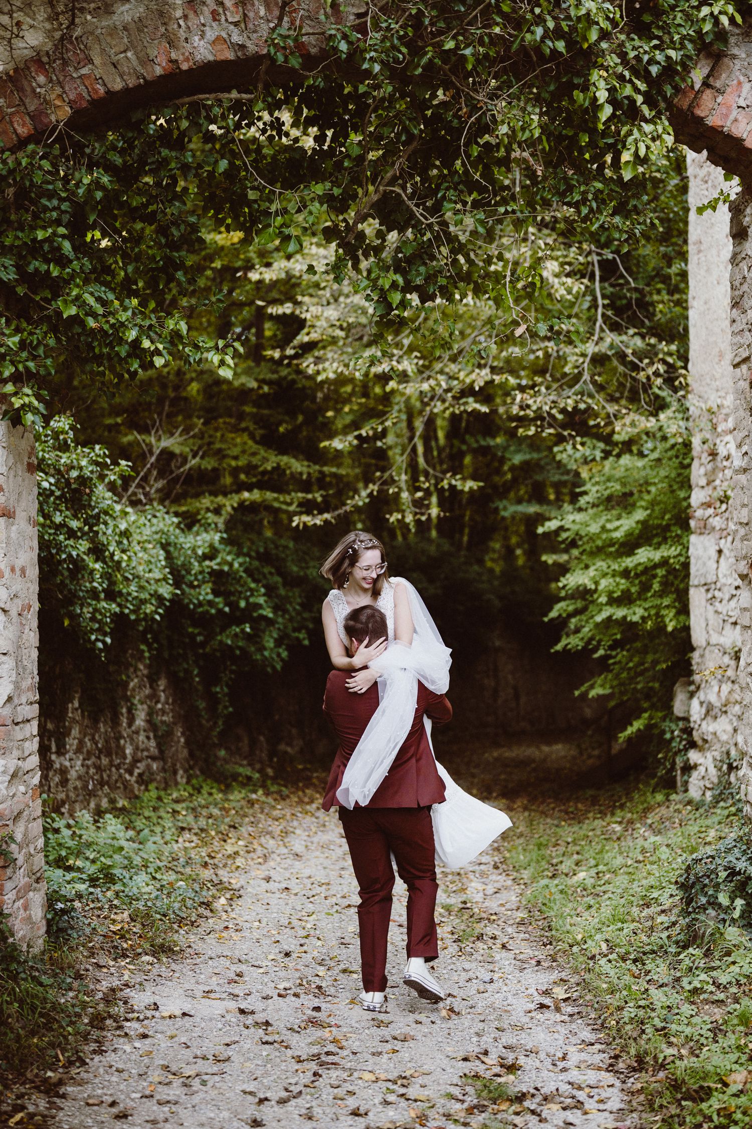 A couple dances playfully together on a leaf-covered path surrounded by lush green trees in a romantic forest setting.