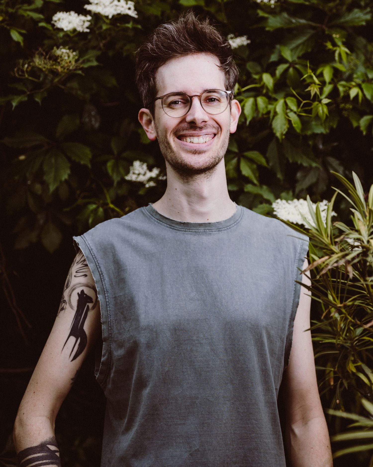 A person in a gray sleeveless shirt smiles while standing in front of green foliage and white flowers.