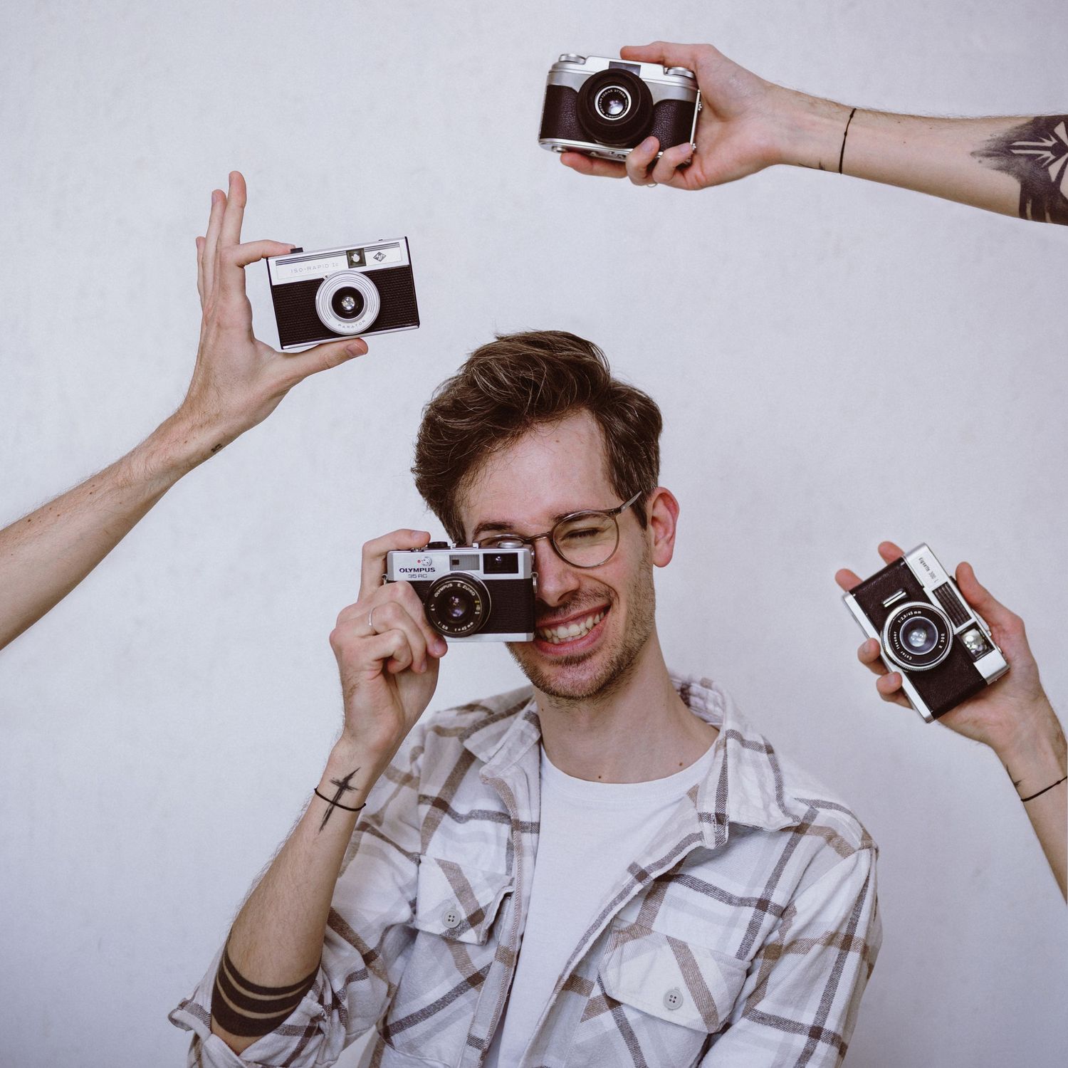 Multiple vintage film cameras surround a person in a plaid shirt laughing against a light background.