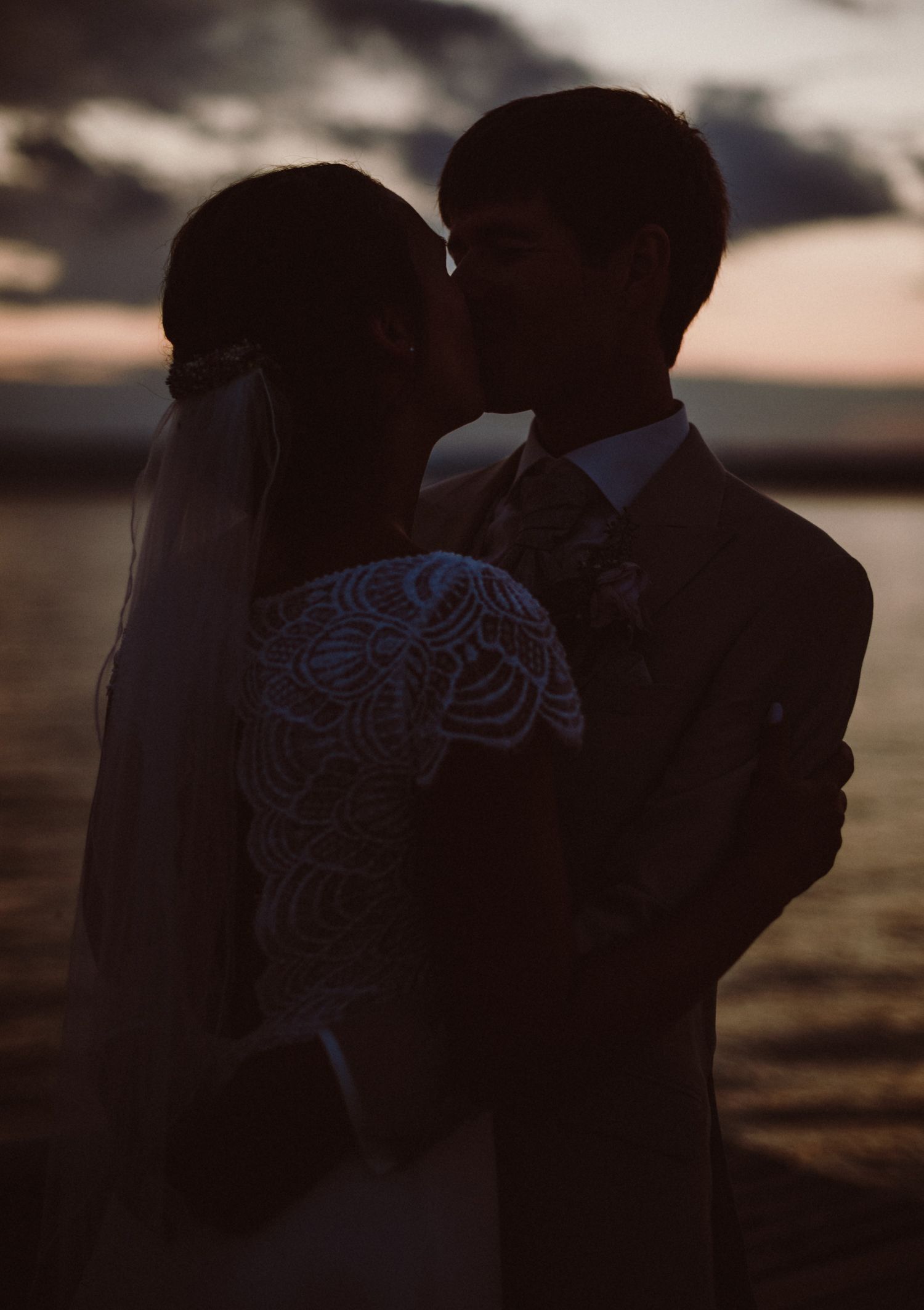 A silhouetted couple in wedding attire share a romantic moment at sunset, with the bride wearing a detailed lace gown.