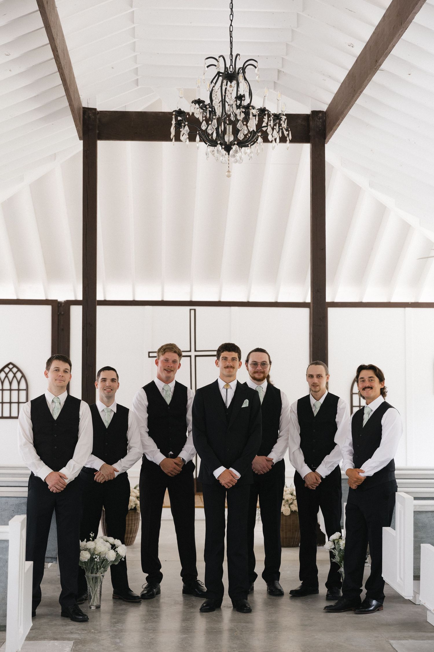 Groom and six groomsmen in black vests and ties pose in white chapel under elegant chandelier.