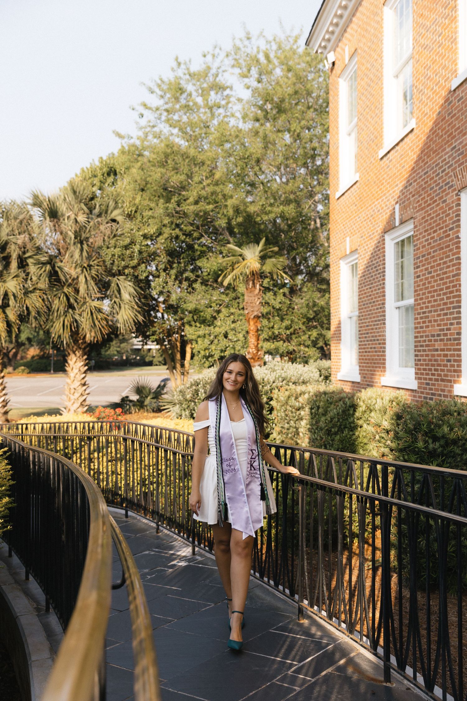 Young woman in white blazer and floral dress walks along a railed balcony beside a brick building with palm trees.