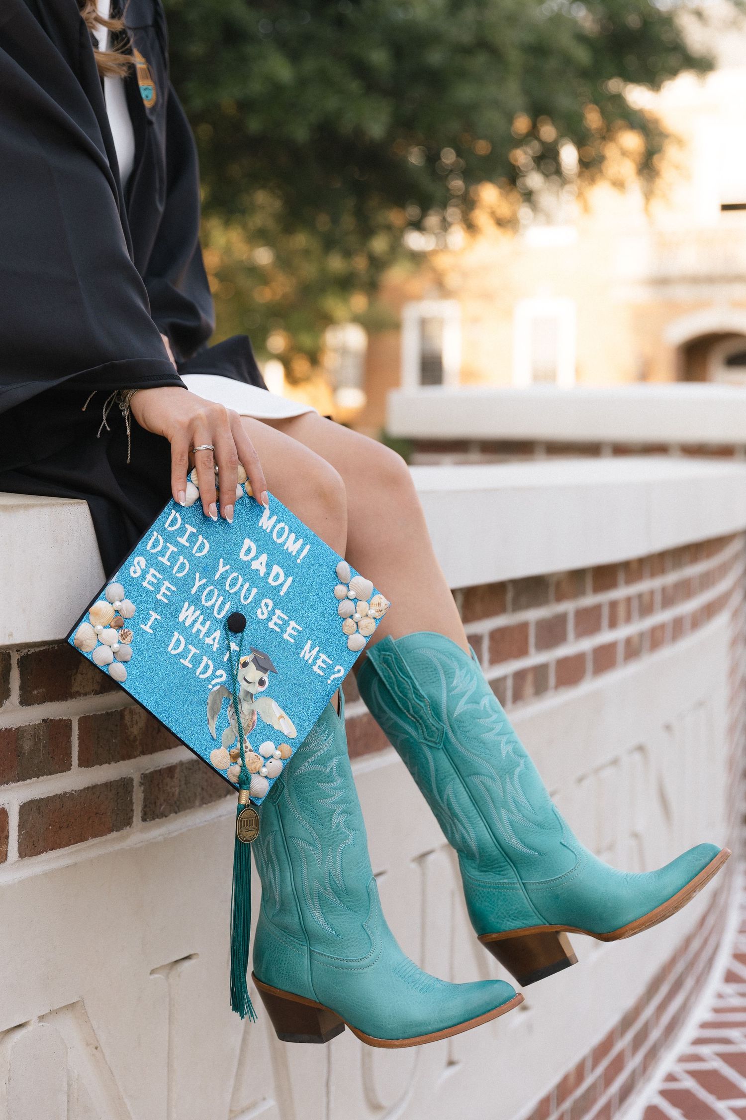 Graduate in teal cowboy boots holds decorated cap reading Mom Dad Did You See Me Did What I Did.