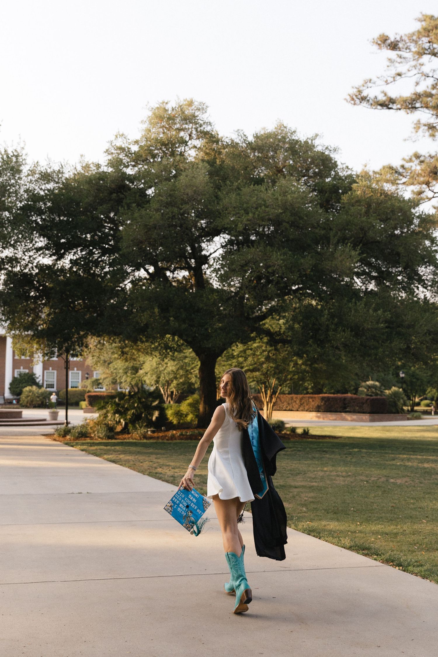 A young woman in a white dress and teal boots walks on a campus path, holding a graduation cap and gown.