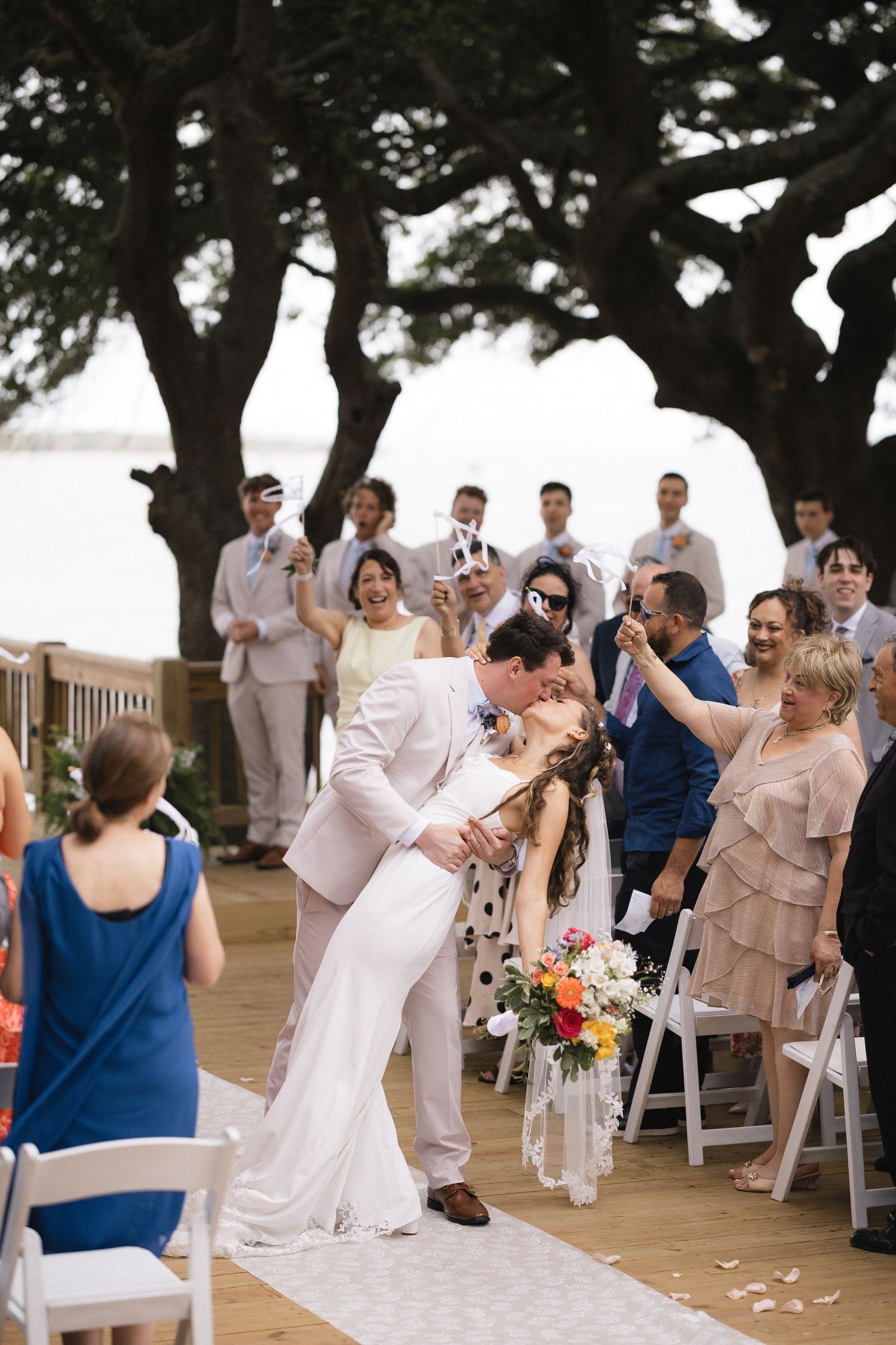 Bride and groom share first kiss at outdoor waterfront wedding ceremony surrounded by guests cheering on decorated deck.