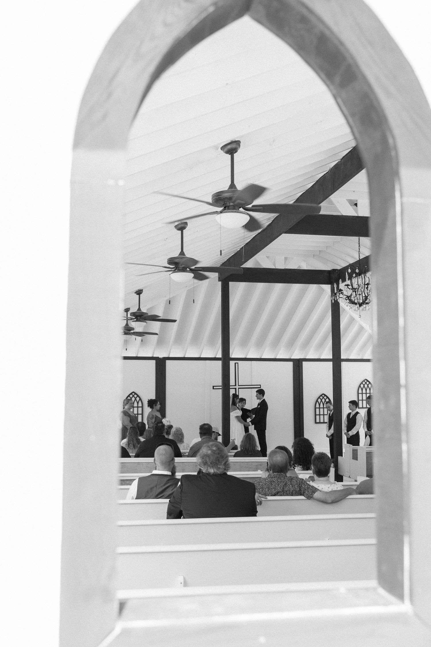 Black and white photo of a wedding ceremony viewed through an arched window, with guests seated in pews.
