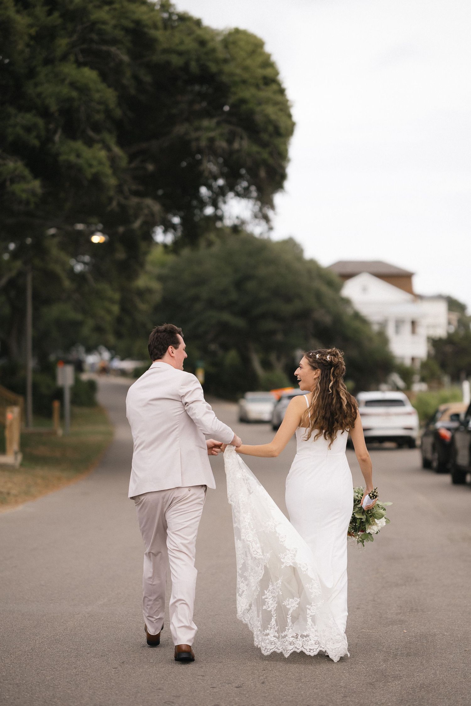 Bride and groom walking hand in hand down a tree-lined street, bride in lace gown holding bouquet.