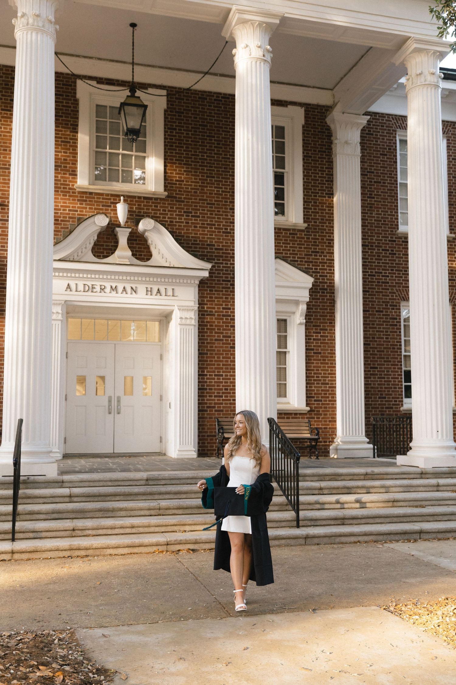 A young woman in a white dress holds a green jacket outside Alderman Hall, a brick building with white columns.