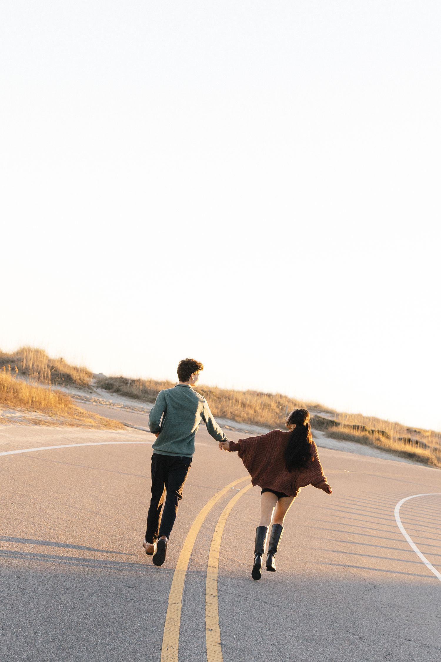 Couple holding hands running down a scenic coastal road at golden hour, viewed from behind.