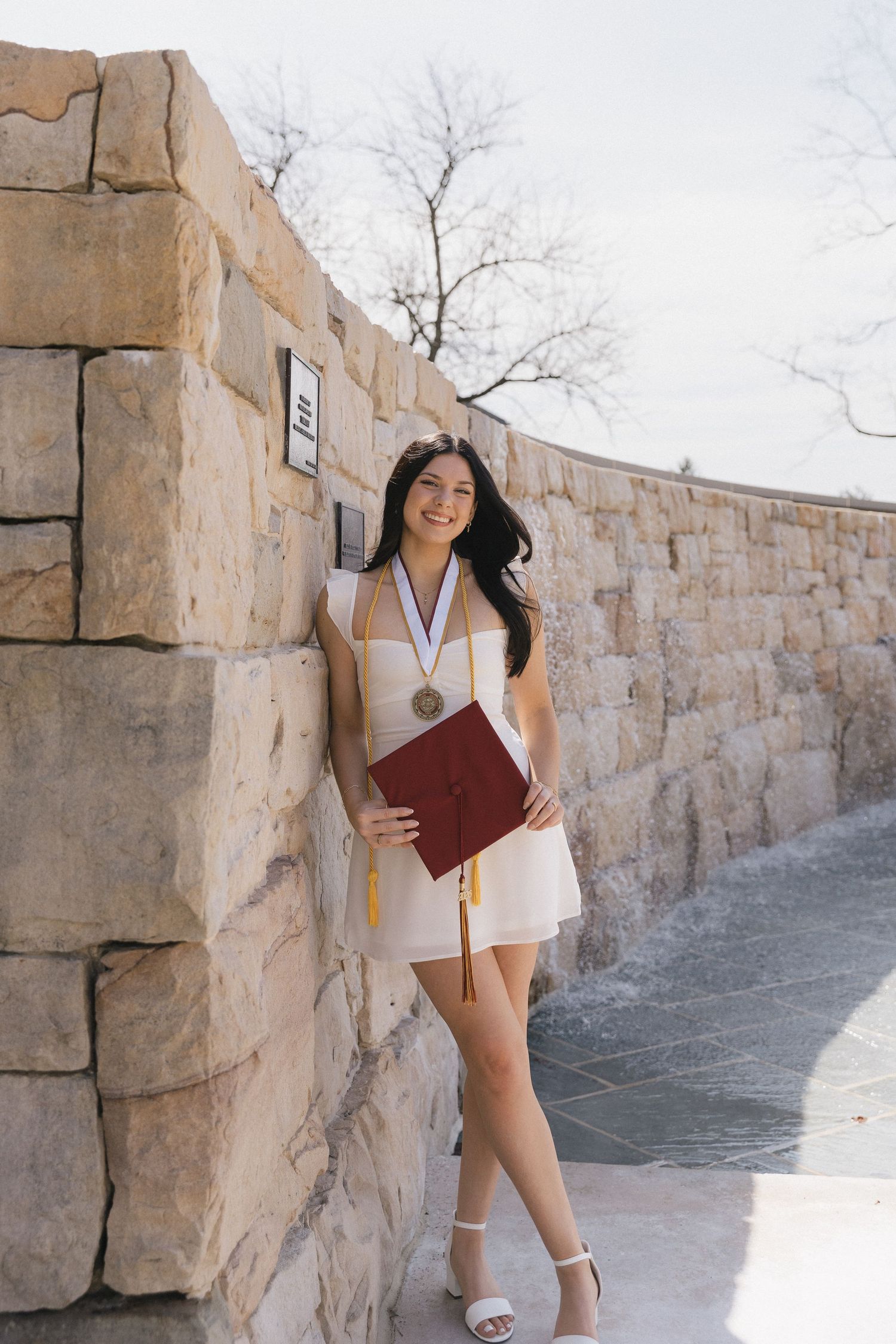 Young woman in white dress holding diploma and wearing medal, posing by stone wall at graduation.