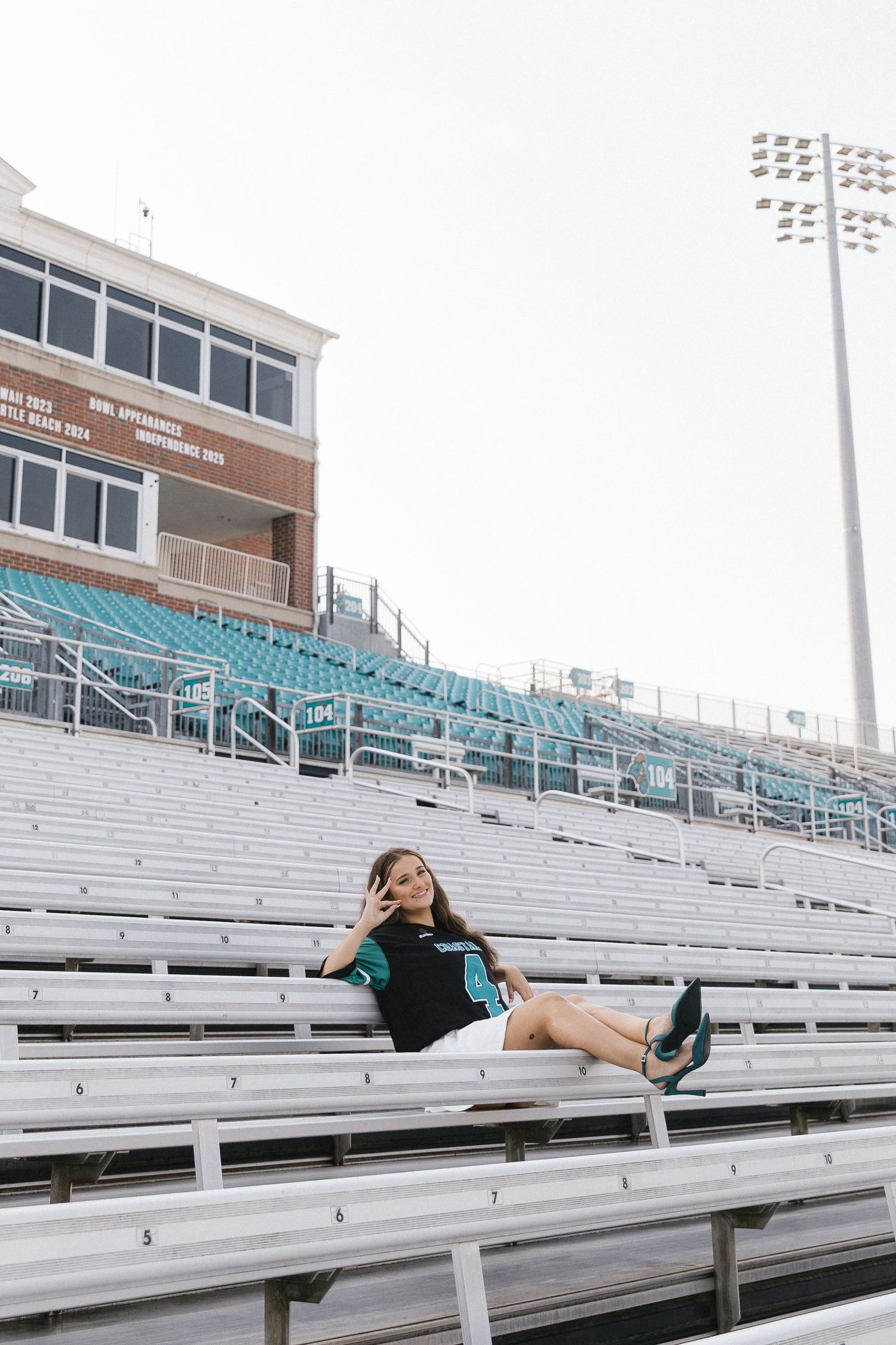 Young woman in black outfit sitting on bleachers at a stadium, posing with a peace sign gesture.