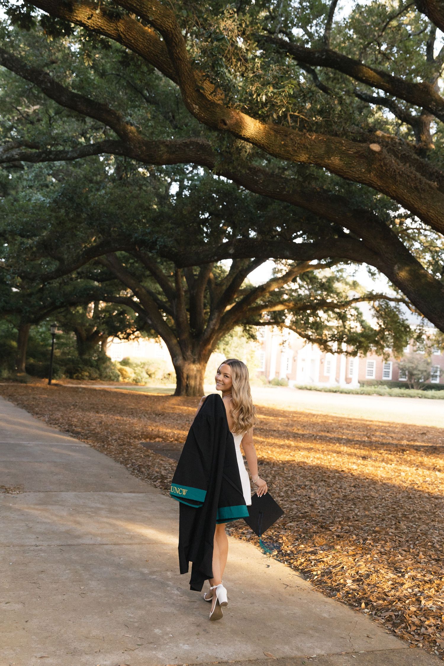 Young woman in graduation gown and cap walks along tree-lined campus path during golden hour.